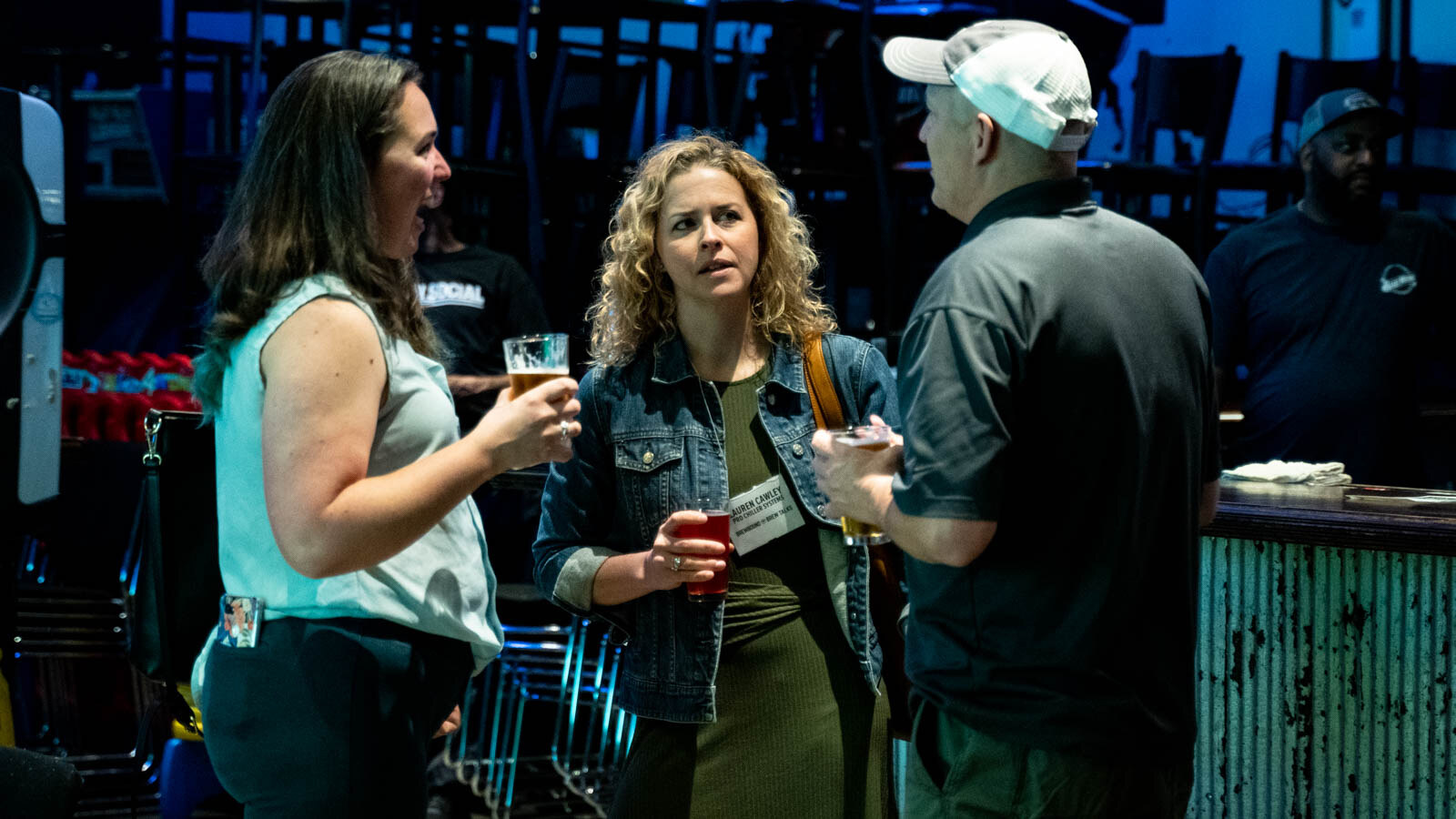 Two women and a man in a ball cap standing at a bar, talking and holding glasses of beer.