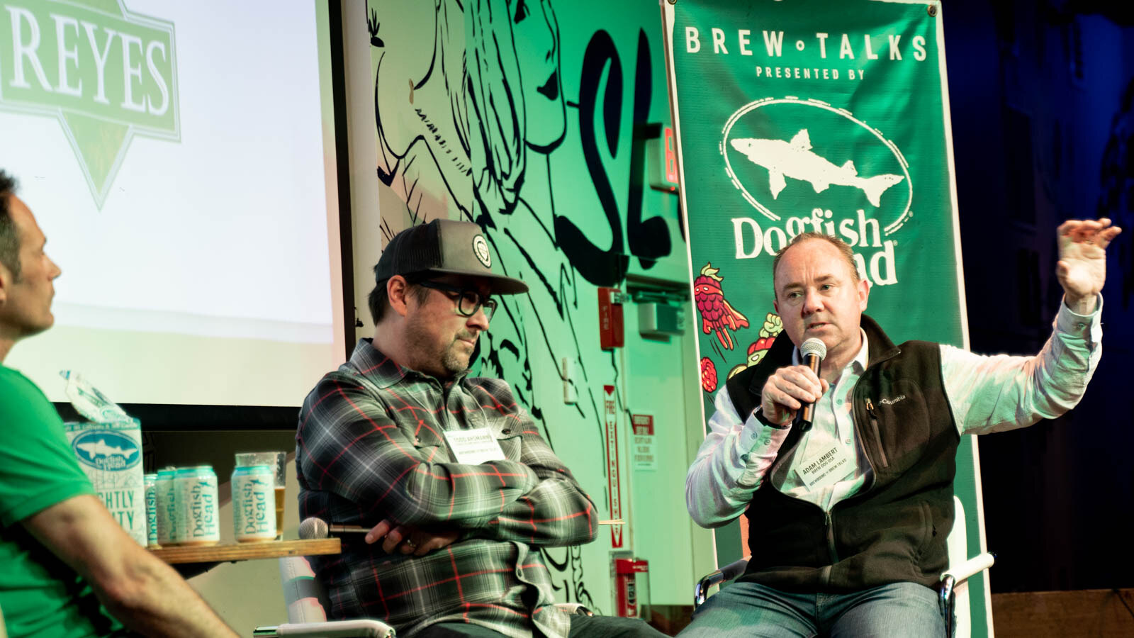 A man with a microphone gesturing mid-sentence next to a man in a plaid shirt, with a green Dogfish Head Brew Talks banner behind them.