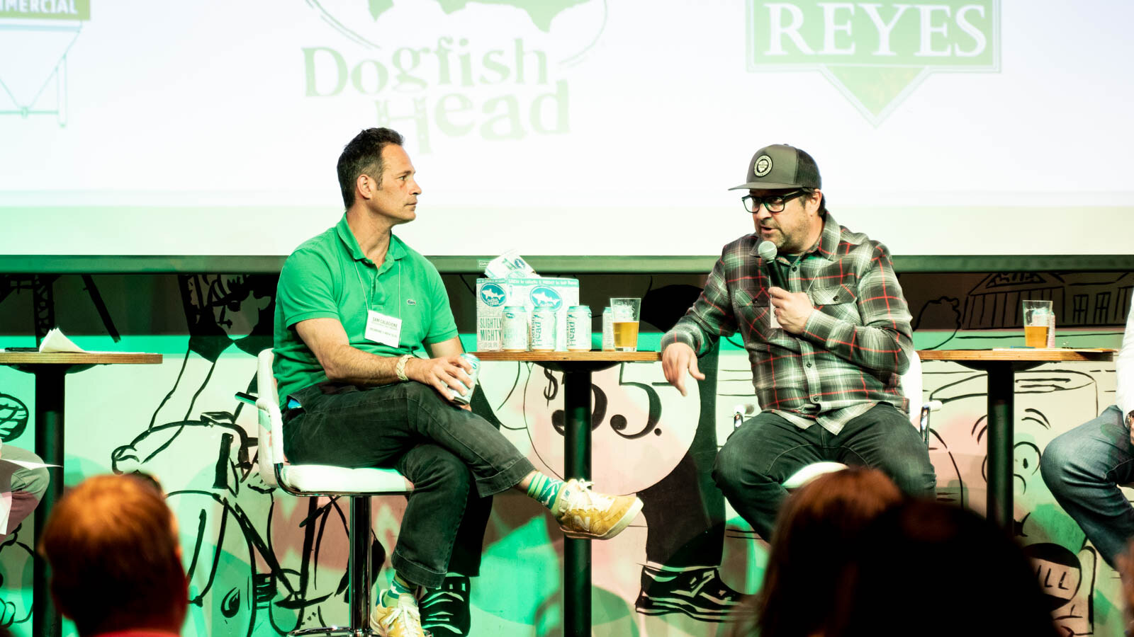 Two men seated on white stools, one in a green polo and one holding a microphone, with Dogfish Head and Reyes logos on the screen behind.