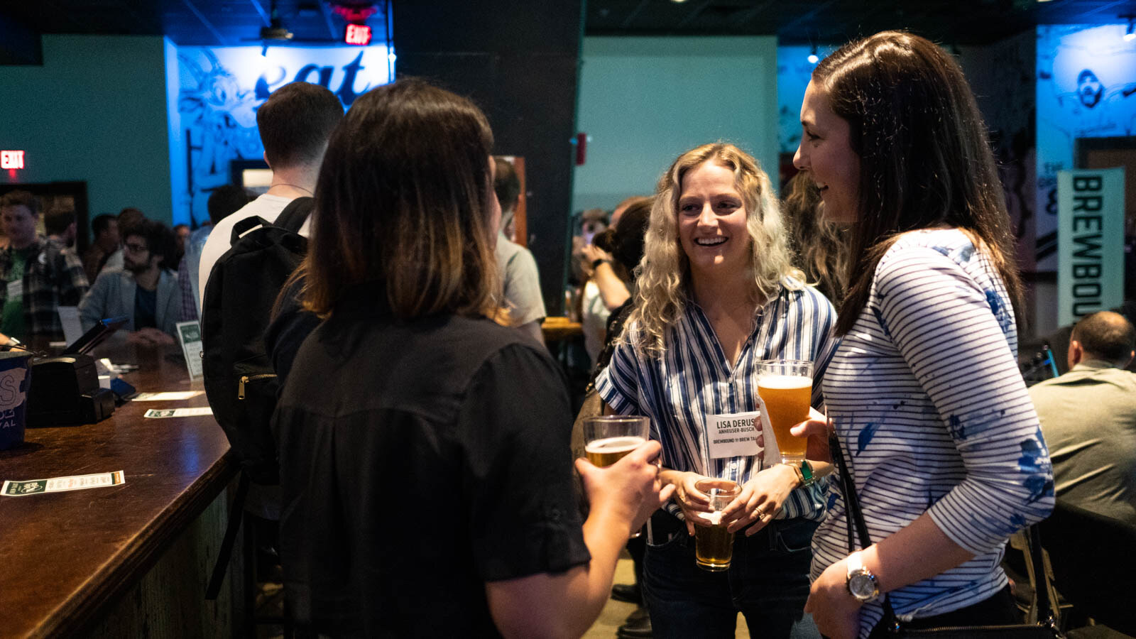 Two women laughing and talking with glasses of beer in a bar, with a Brewbound banner at the right edge.