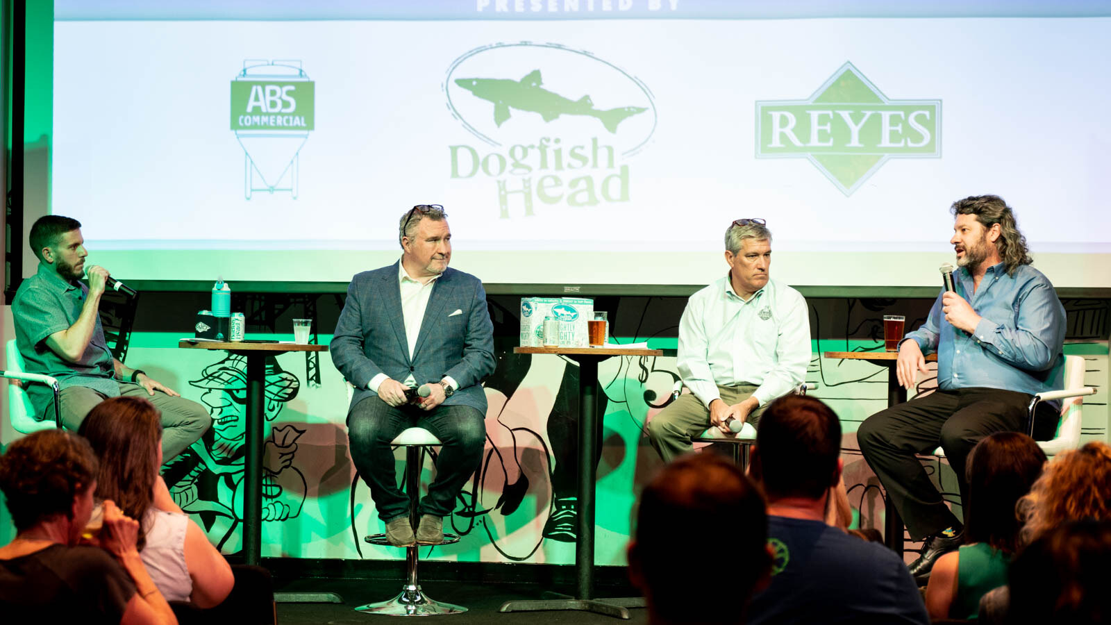 Four people seated on stools in a row on stage, the moderator holding a microphone, with sponsor logos on the screen behind them.