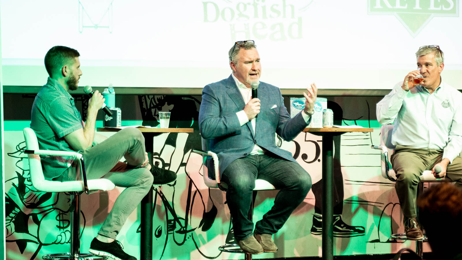A man in a blue blazer speaking into a microphone between a moderator and a man drinking a beer, seated on stage.