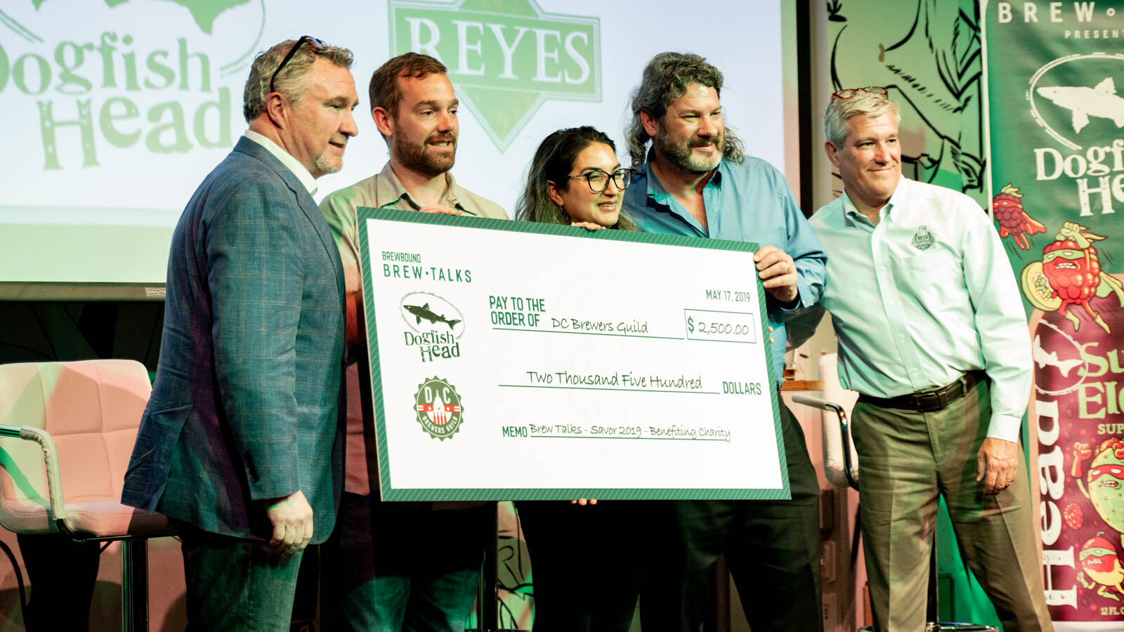 Five people on stage holding a large check payable to the DC Brewers Guild for $2,500, with a Brew Talks banner behind them.