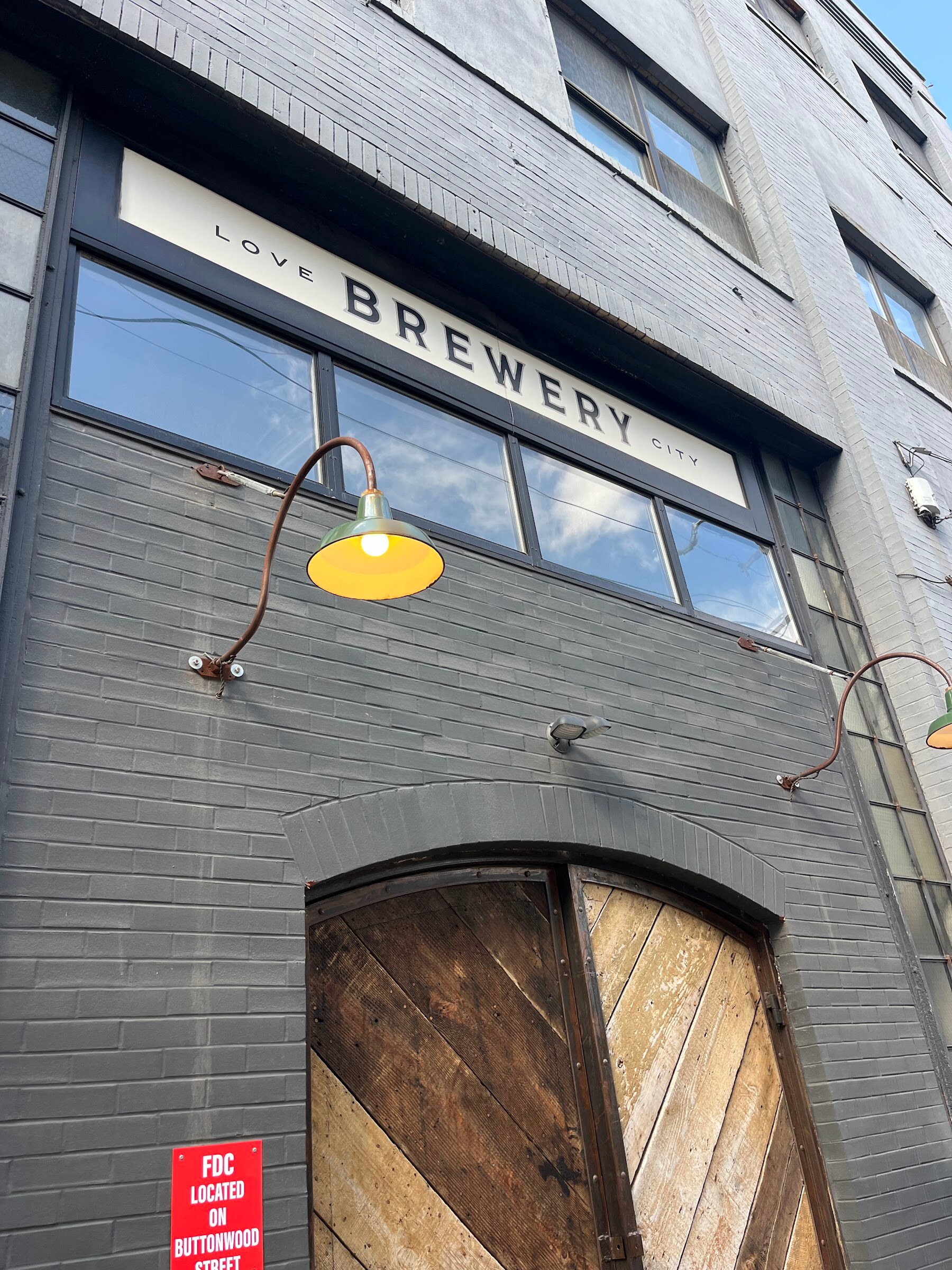 Gray brick exterior of Love City Brewery with a wooden arched door and a gooseneck lamp.
