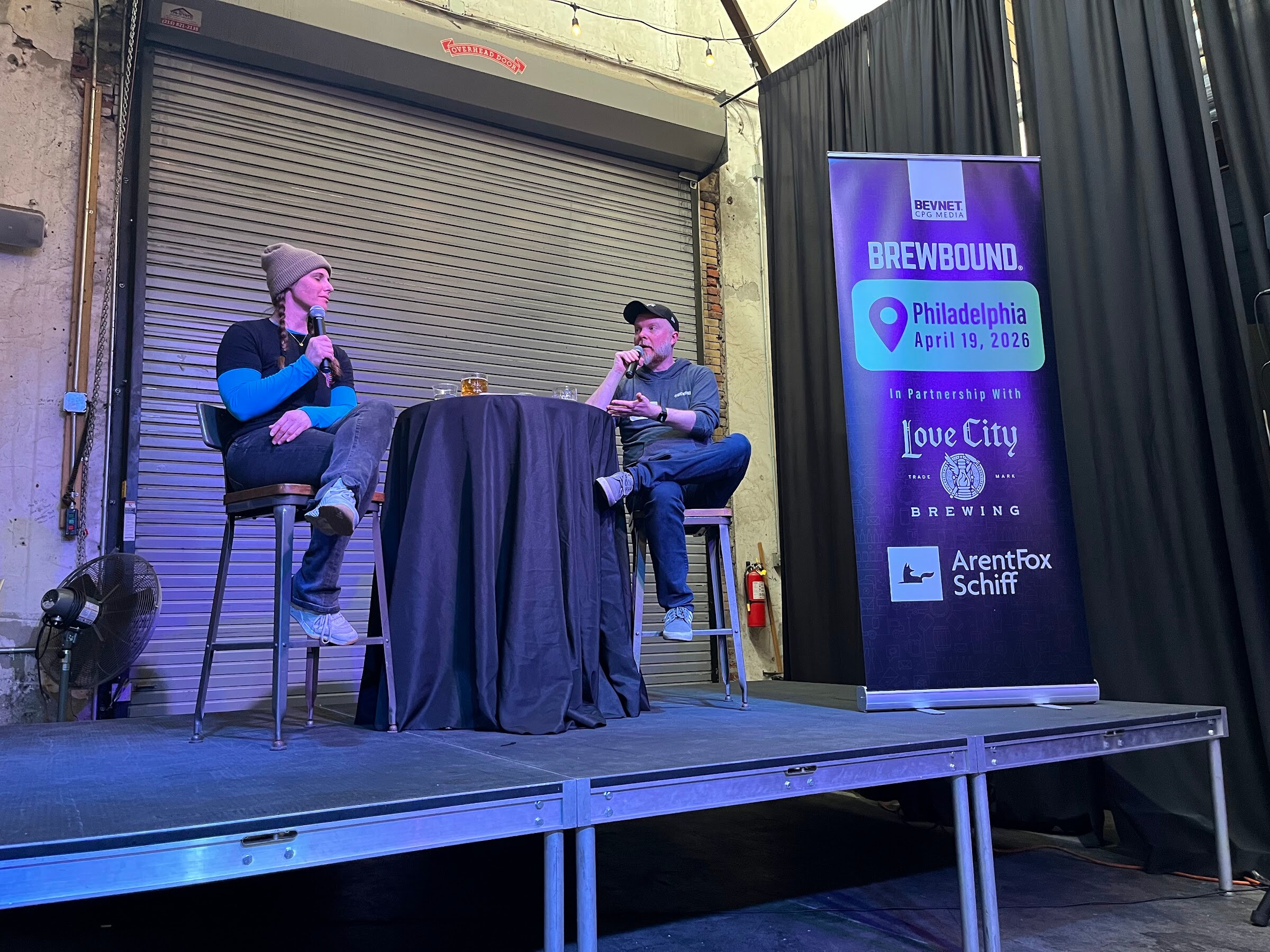 Two people on stools talk into microphones on a small stage beside a Brewbound banner for Philadelphia, April 19, 2026.