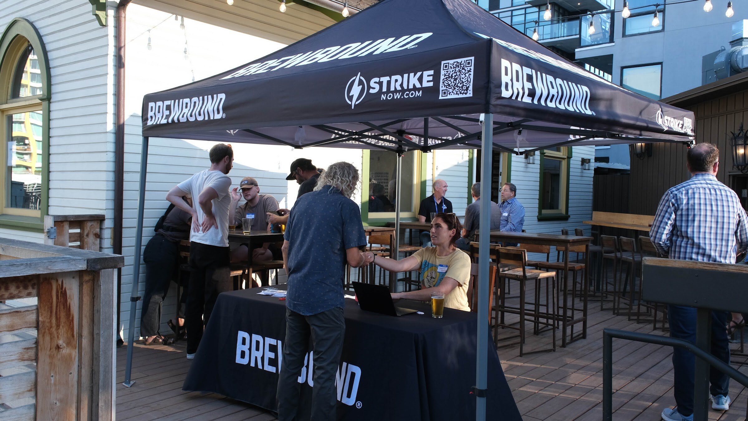 Black Brewbound canopy and table on a wooden patio where attendees check in at a laptop, with guests standing and sitting around tall tables.
