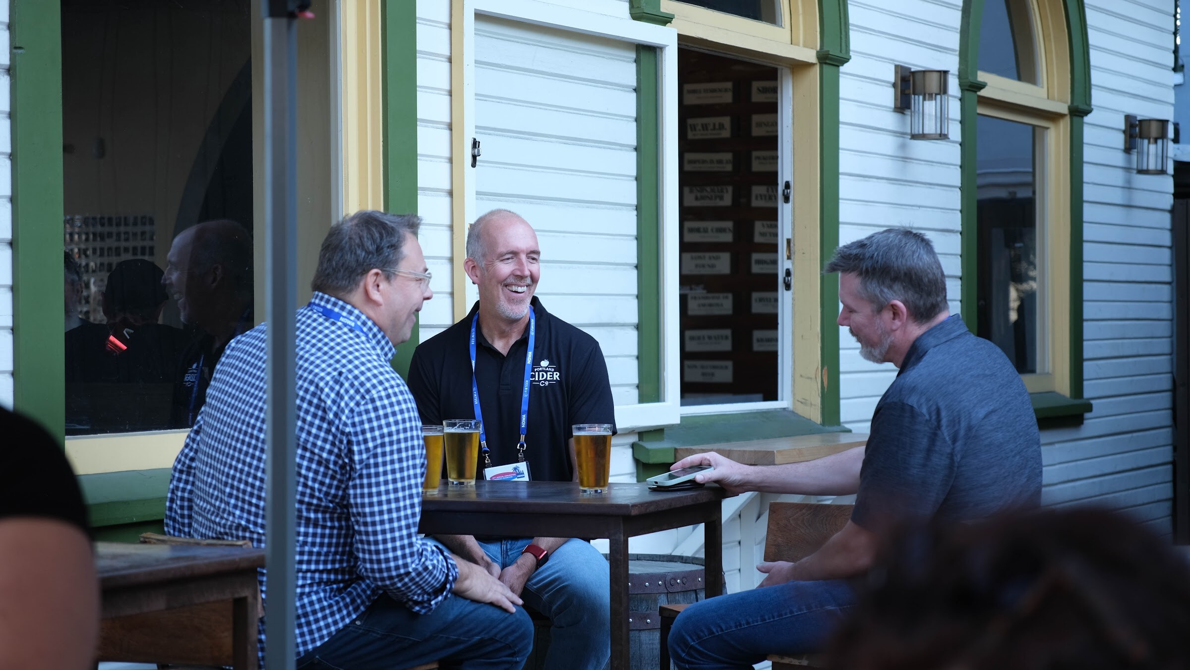Three men seated at an outdoor table with glasses of beer, laughing, with white clapboard walls and a green window frame behind them.