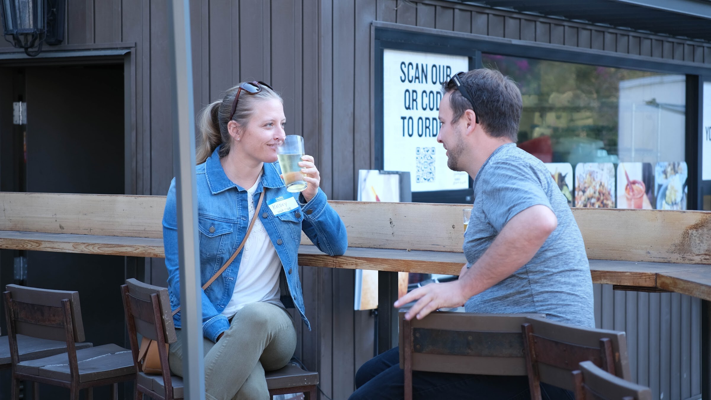 Woman in a denim jacket holding a beer and a man in a gray shirt talking at a wooden rail on an outdoor patio.