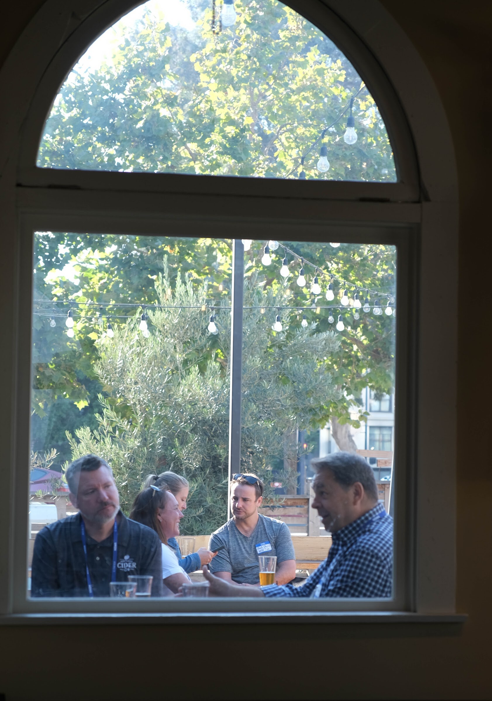 Arched window looking out on trees and string lights, with four people seated at a patio table in the distance.