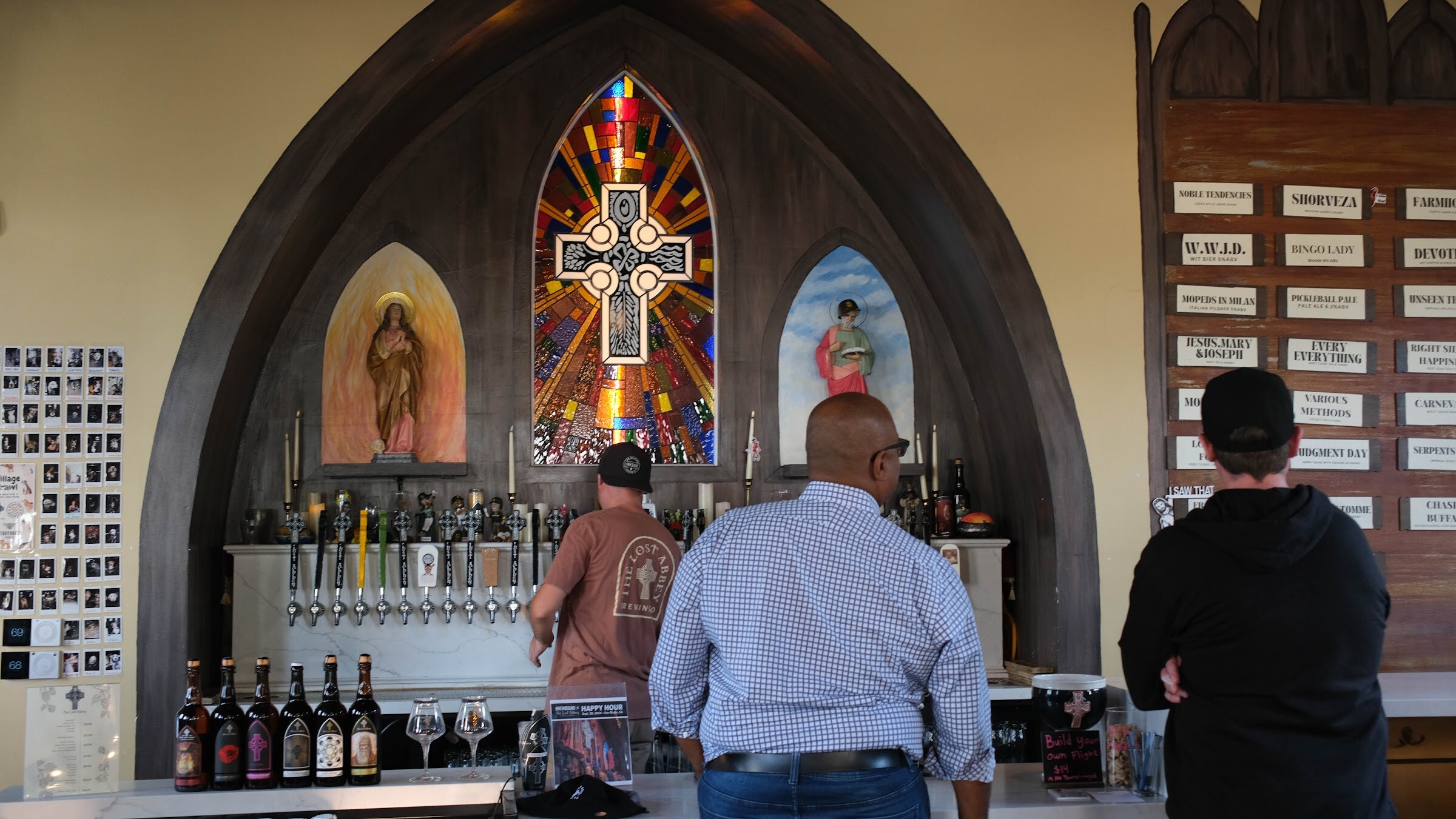 Man in a plaid shirt and others at the long bar with rows of tap handles and bottles beneath a stained glass window.