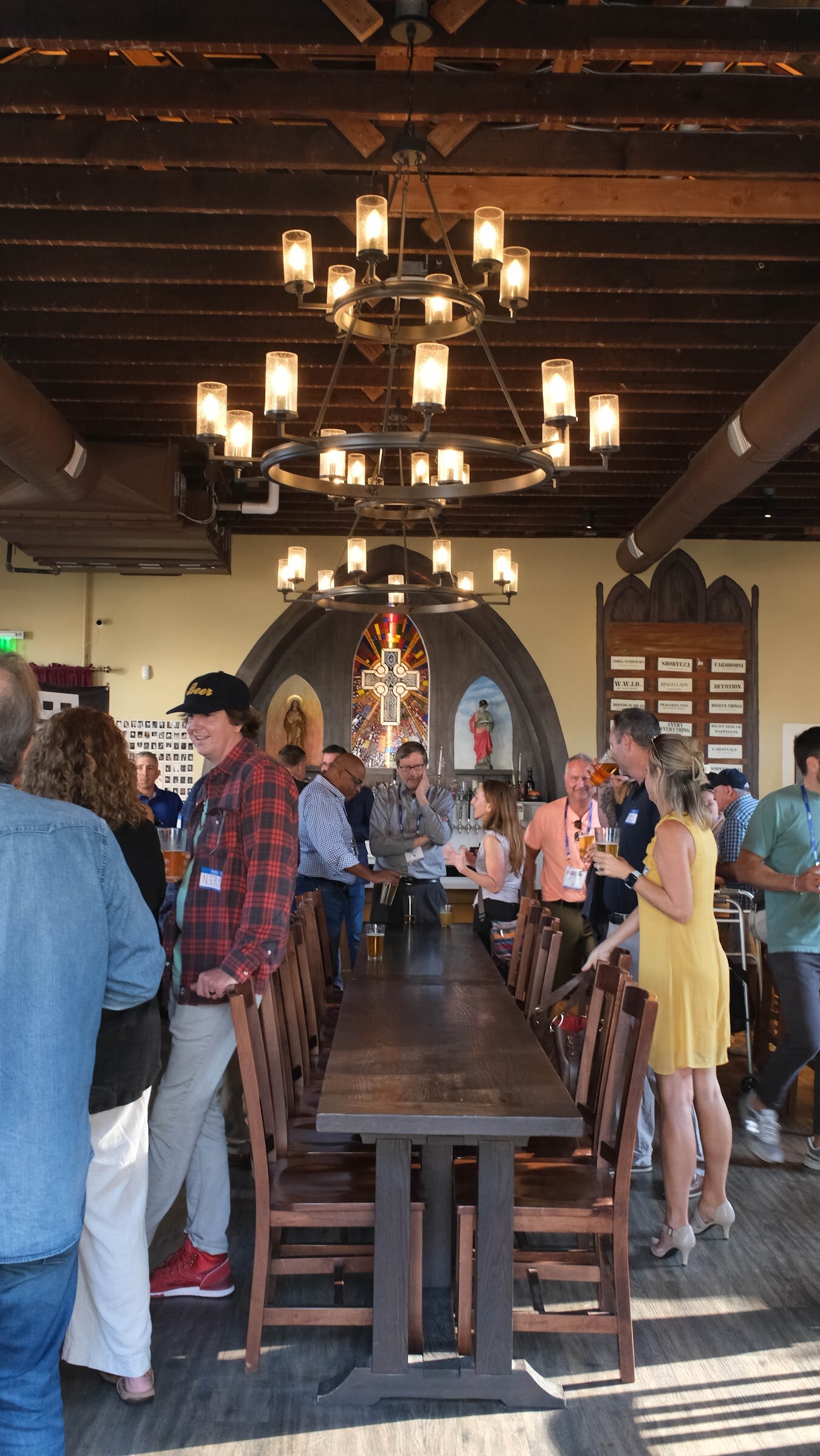 Long wooden table under iron chandeliers with attendees standing and talking toward the bar and stained glass at the far end.