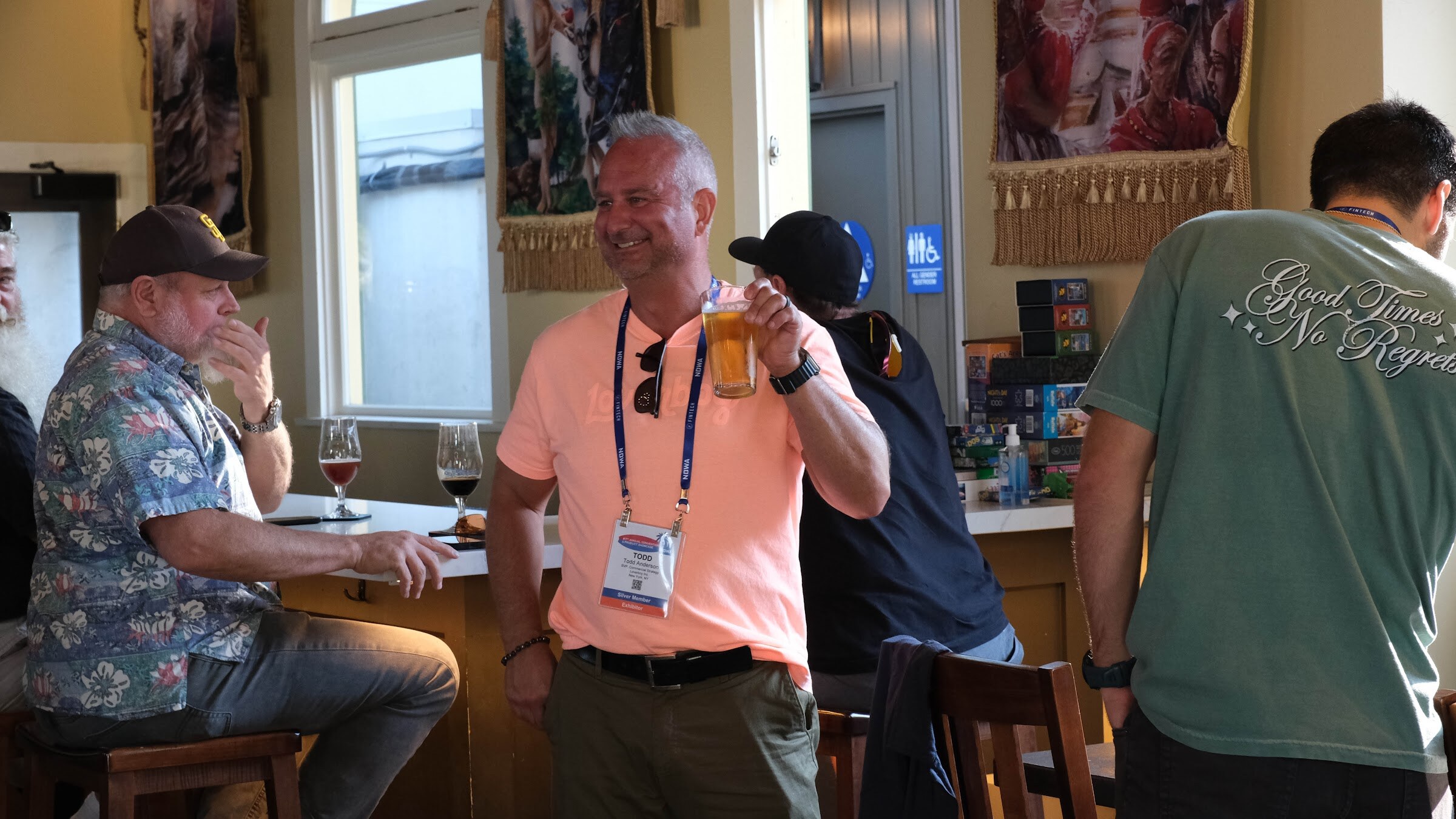 Man in a coral shirt with a conference lanyard raising a beer glass beside other guests at the bar counter.