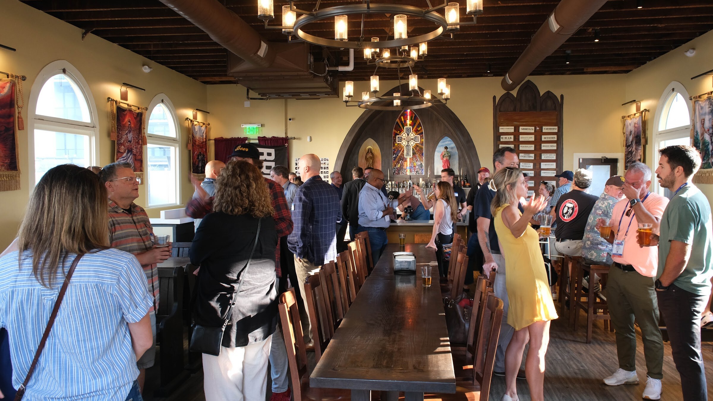 Wide interior view of a church-style taproom with a long table and many guests standing and talking under chandeliers.