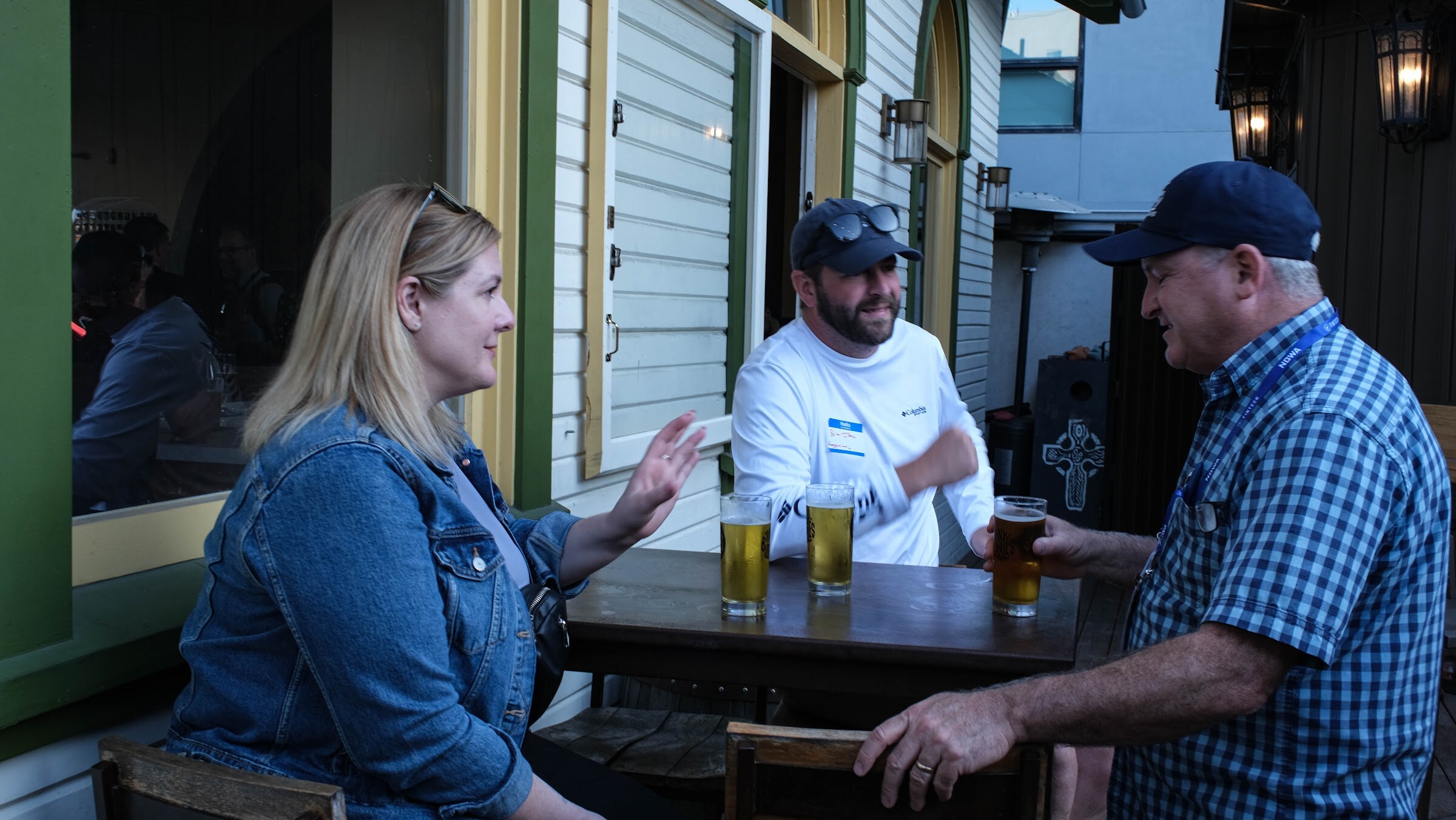 Woman in a denim jacket, a bearded man in a white shirt and a man in a plaid shirt and cap talking at a patio table with three pints of beer.