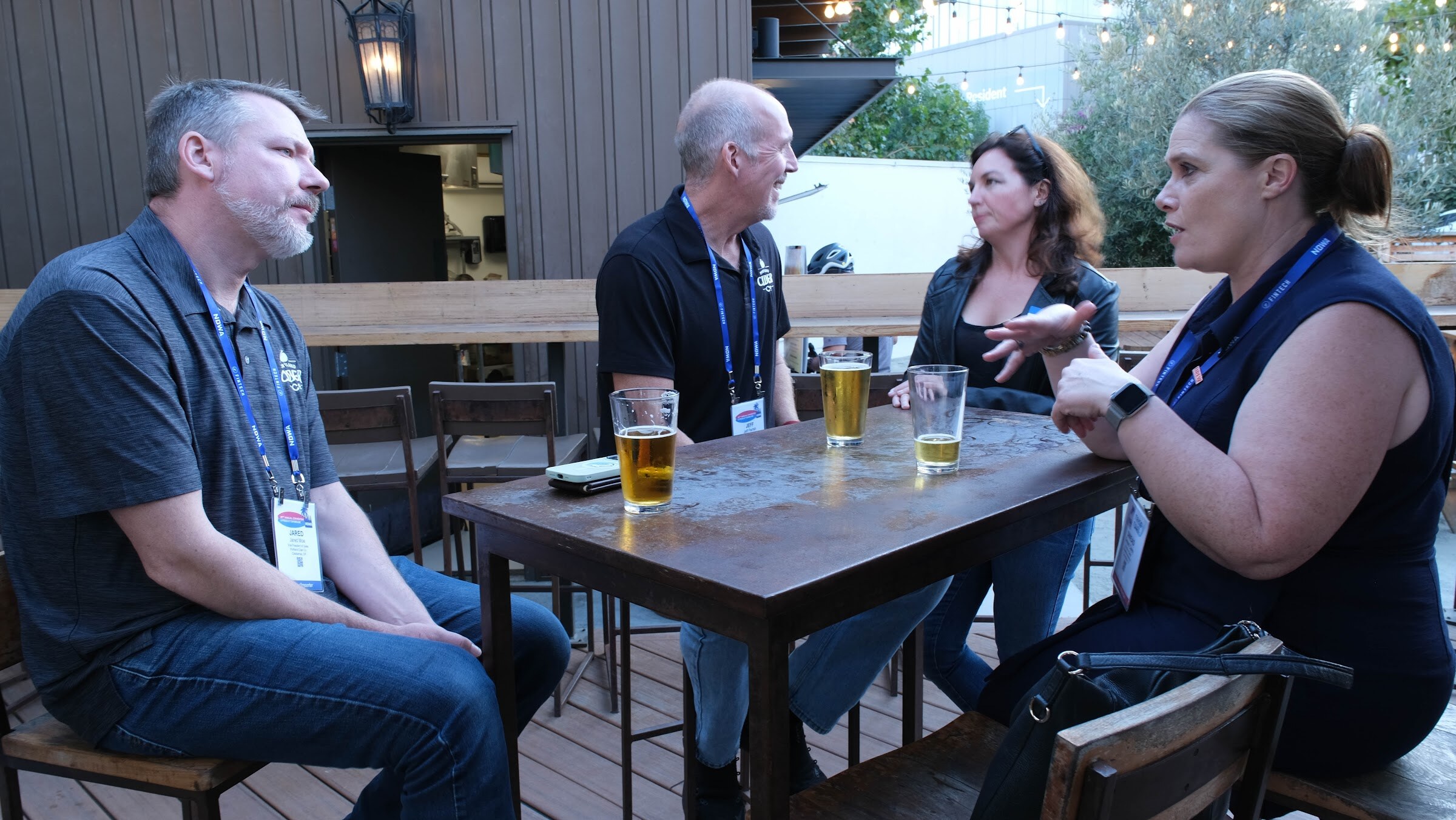 Four people with conference lanyards seated around a table on a patio, with two glasses of beer on the table.