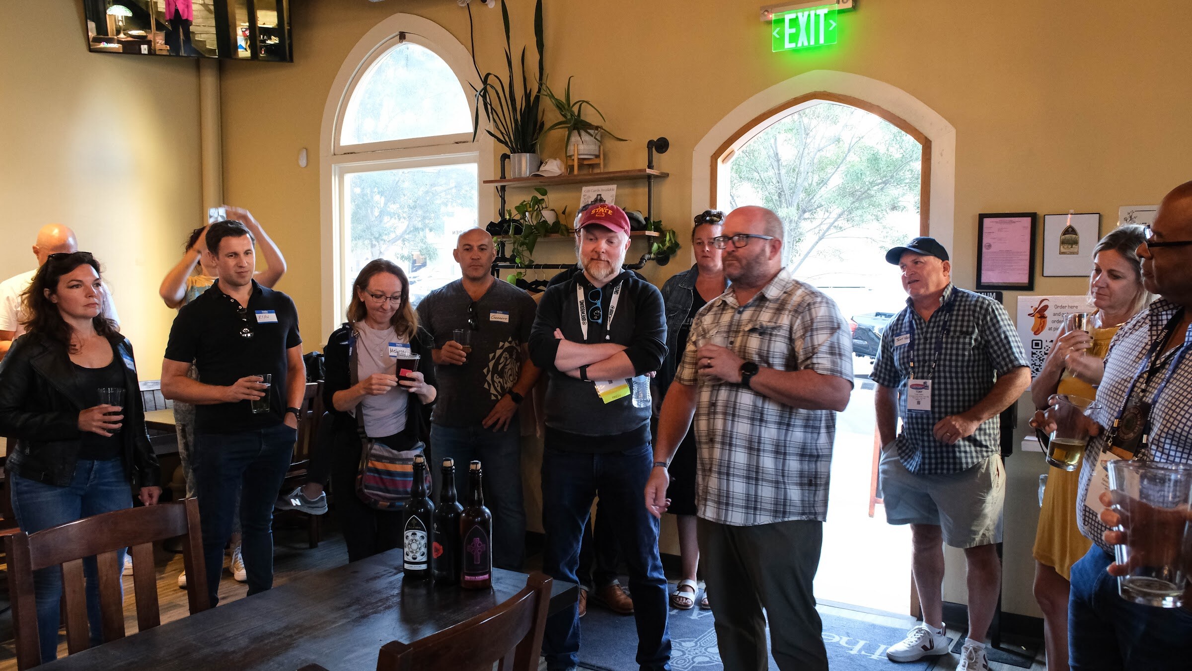 Group of attendees standing close together indoors, holding drinks and listening, with bottles on a table in front.