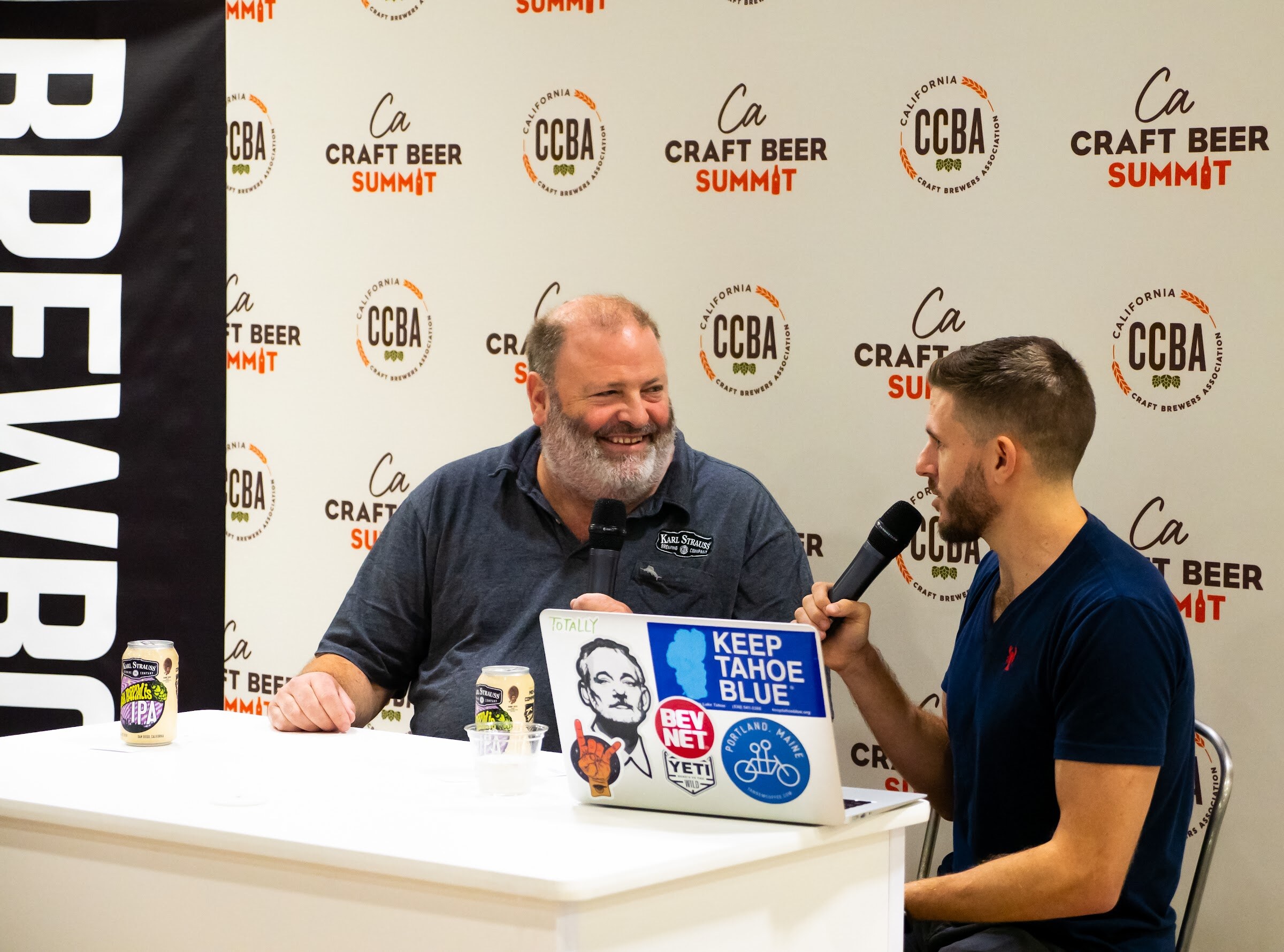 A bearded man in a Karl Strauss shirt smiles into a microphone across a white table from an interviewer, with a laptop covered in stickers and beer cans on the table.