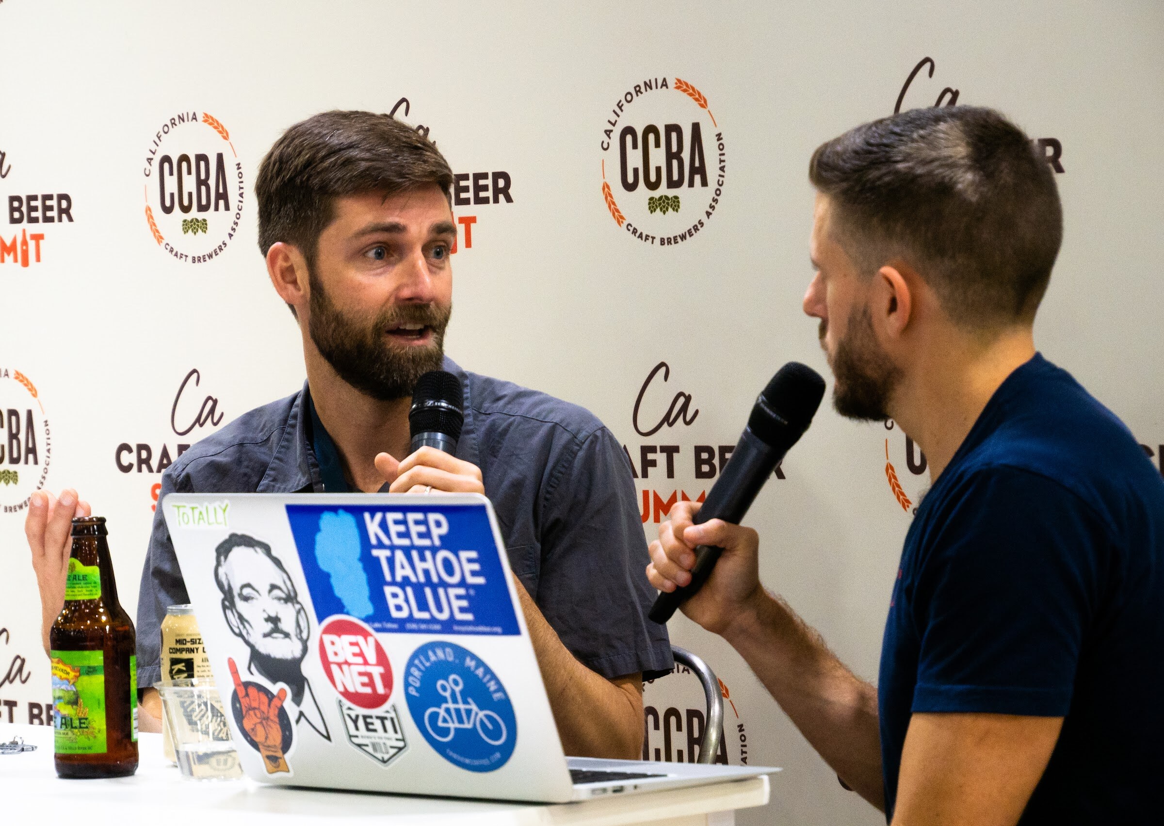 A bearded man in a gray shirt speaks into a microphone across from an interviewer with a laptop covered in stickers reading Keep Tahoe Blue and BevNET.