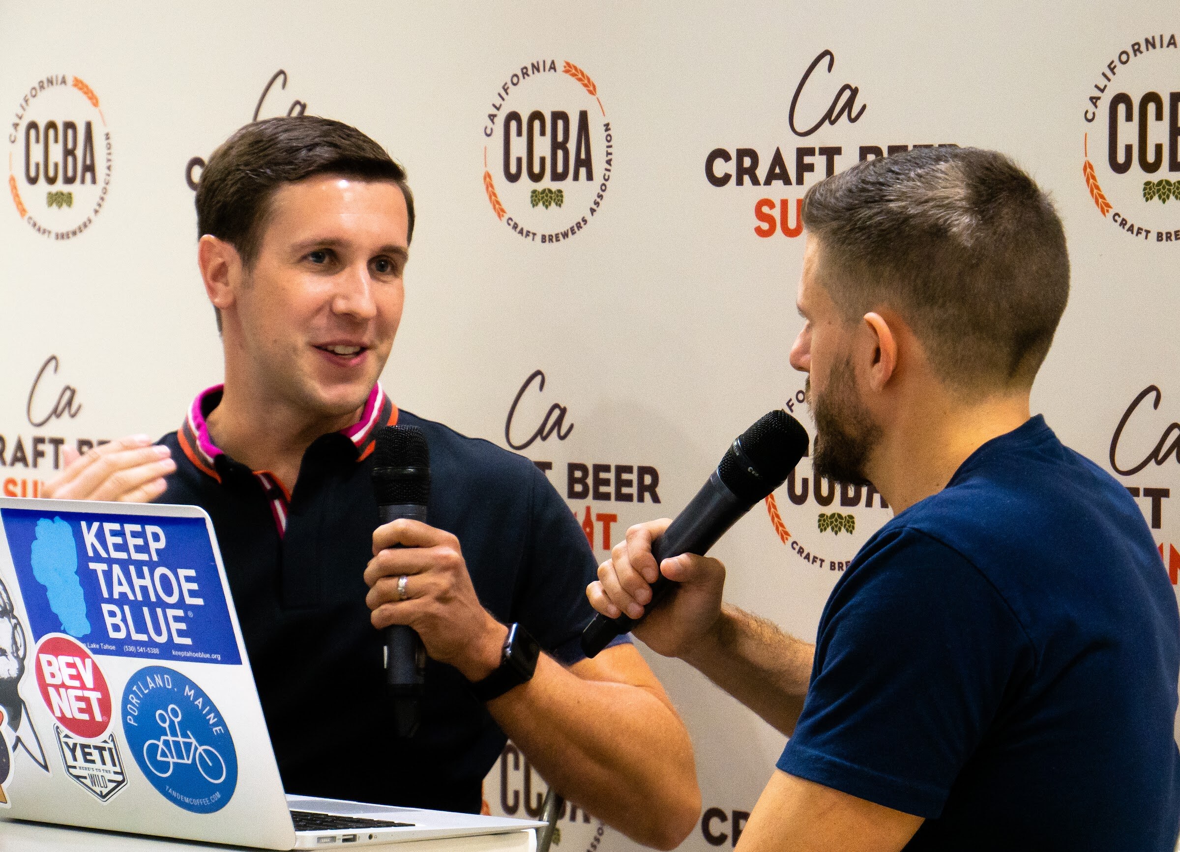 A smiling man in a navy polo holds a microphone in front of a CCBA California Craft Beer Summit backdrop, with a stickered laptop in the foreground.