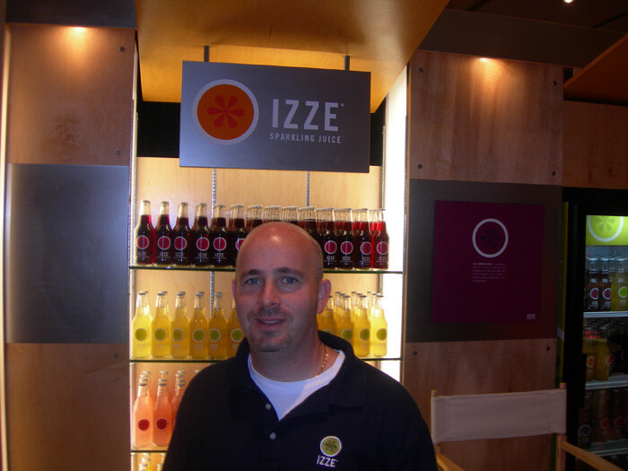 A bald man with a goatee in a black Izze polo stands in front of an Izze Sparkling Juice sign and lit shelves of bottles in red, yellow and pink.