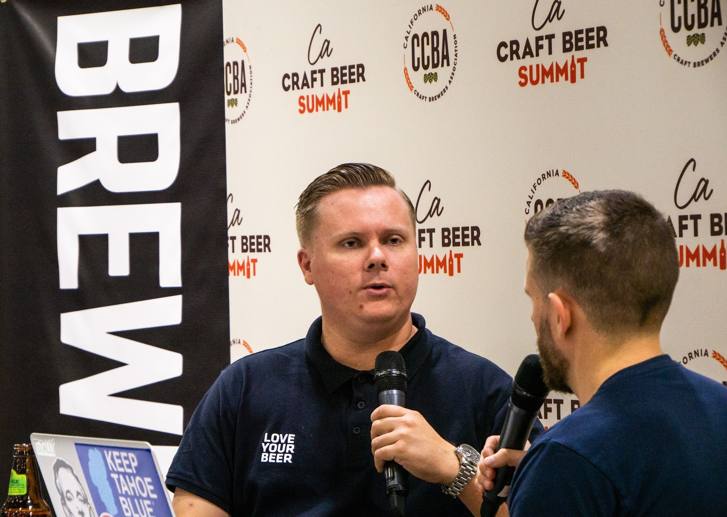 A man in a navy Love Your Beer polo with a watch speaks into a microphone in front of a Brewbound banner and the Craft Beer Summit backdrop.