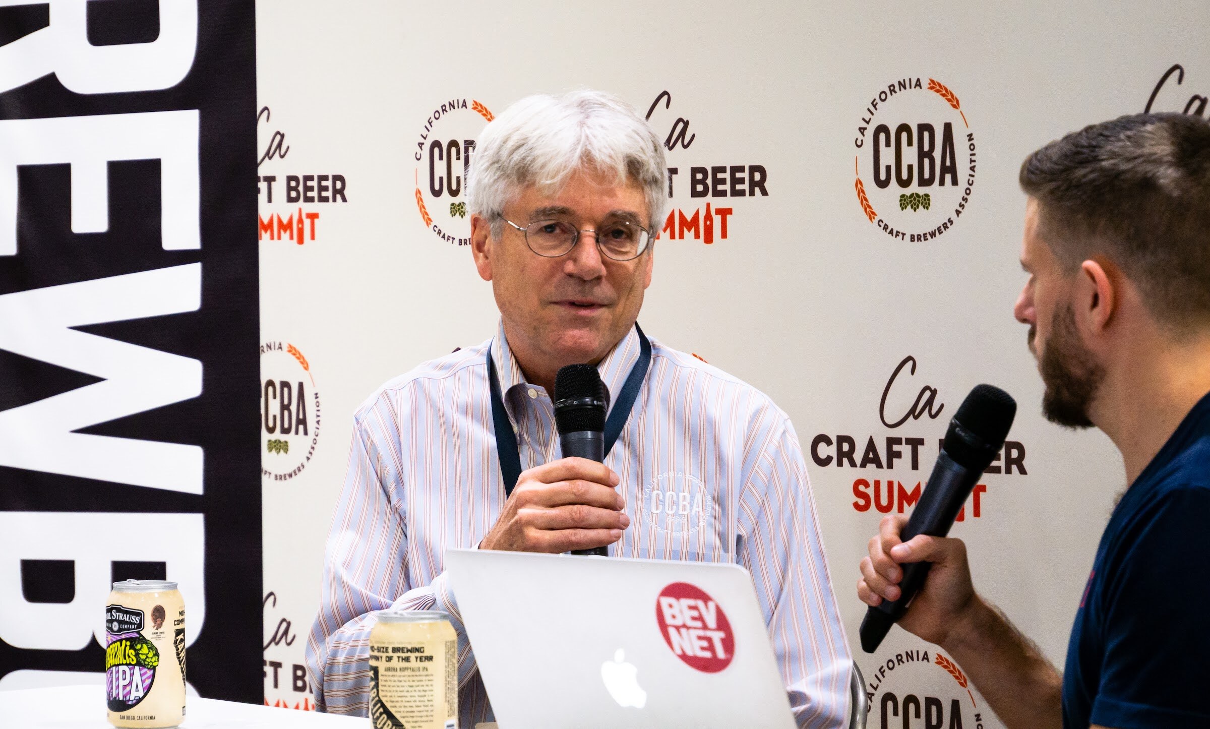 A man in a pinstripe shirt with a CCBA logo and a lanyard holds a microphone beside a laptop with a BevNET sticker and two cans of beer.