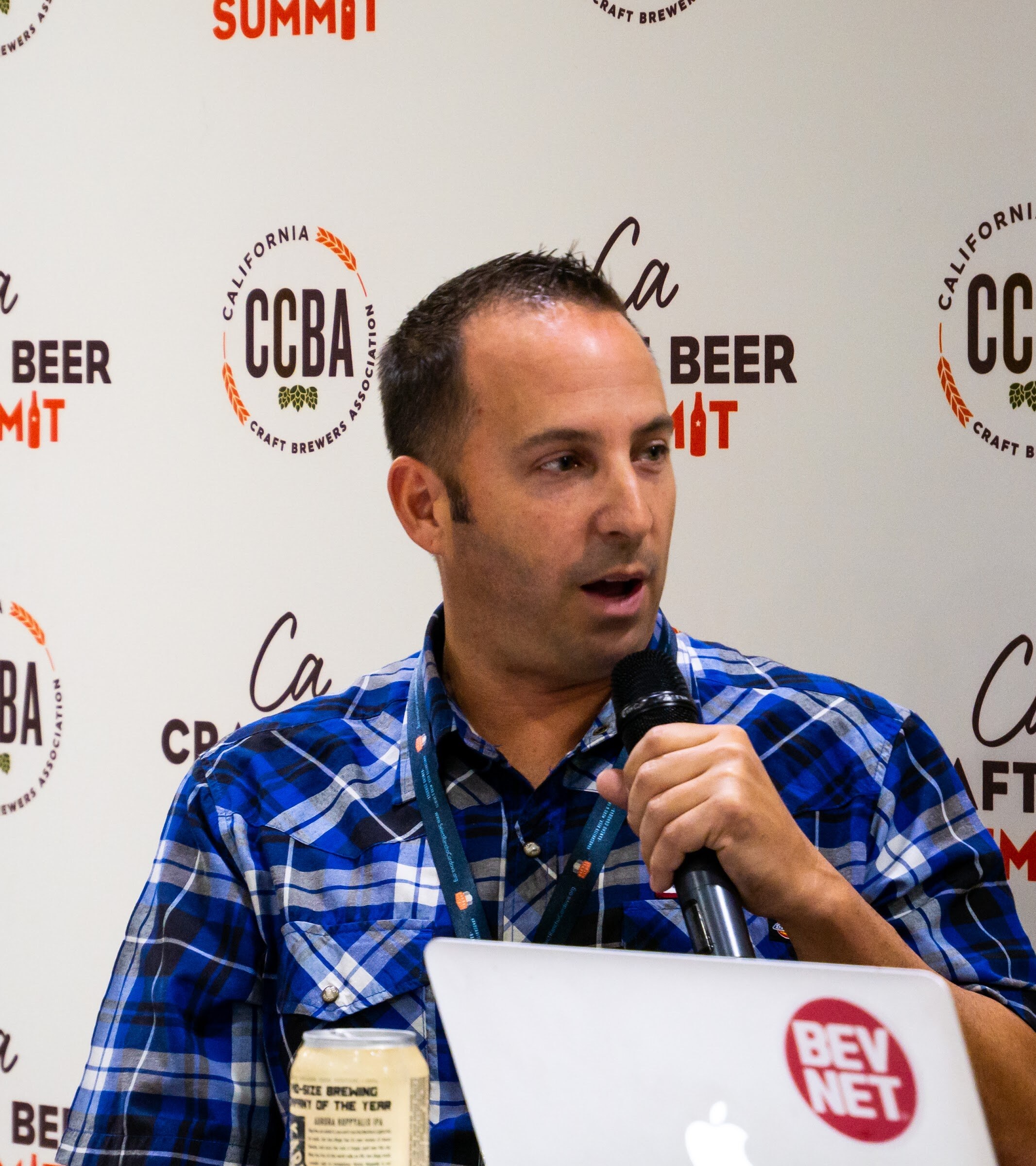 A man in a blue plaid snap shirt with a lanyard speaks into a microphone in front of the CCBA backdrop, with a BevNET laptop and a can of beer.