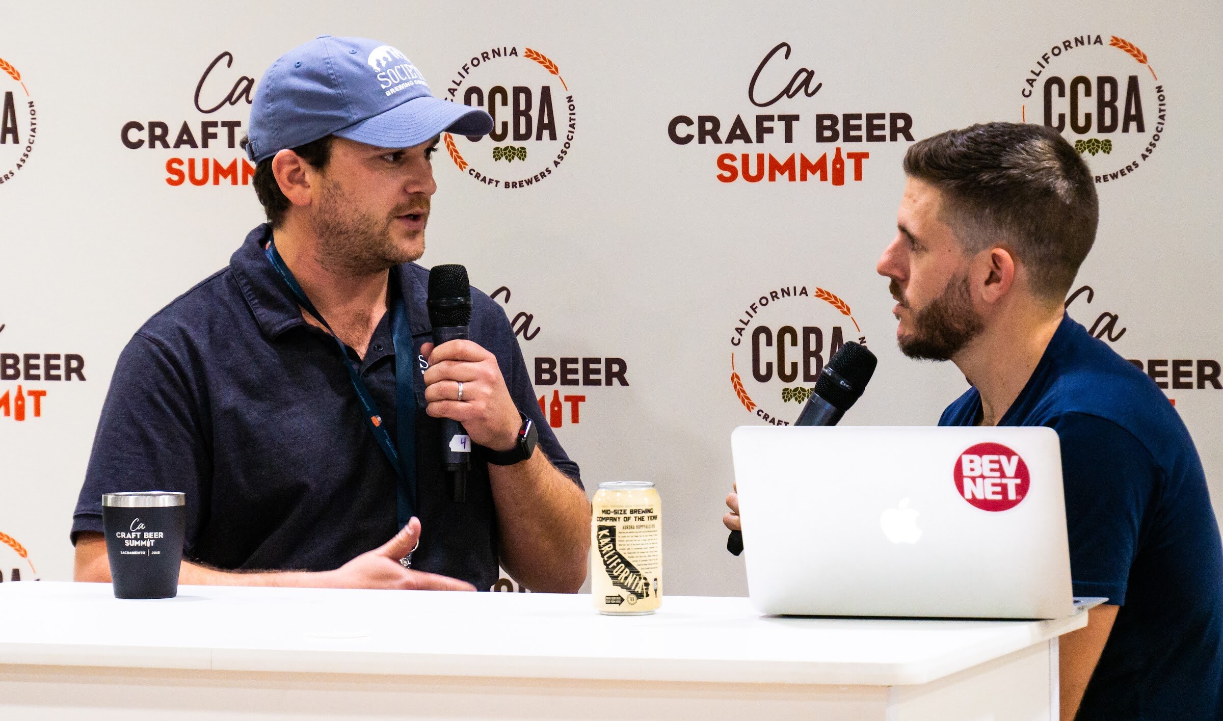 A man in a blue cap and a dark polo with a lanyard speaks into a microphone at a white table, with a California Craft Beer Summit tumbler, a can of beer and a BevNET laptop on the table.