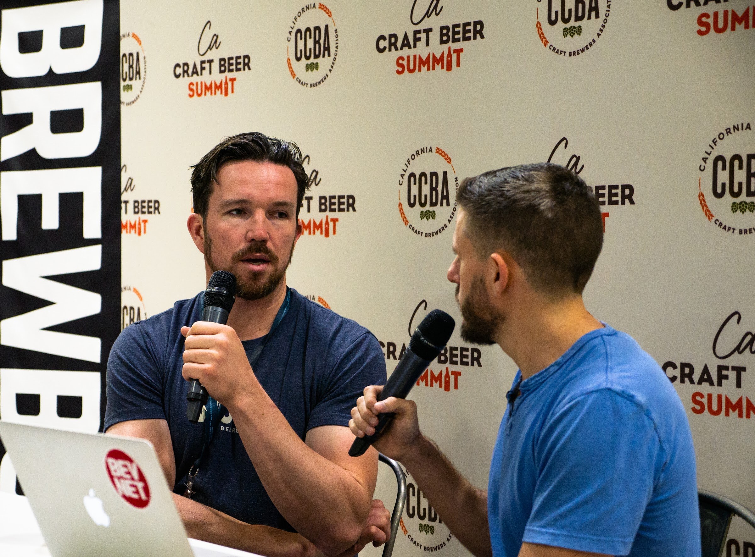 A man with a goatee in a dark T-shirt holds a microphone beside a laptop with a BevNET sticker, facing an interviewer in a blue shirt.