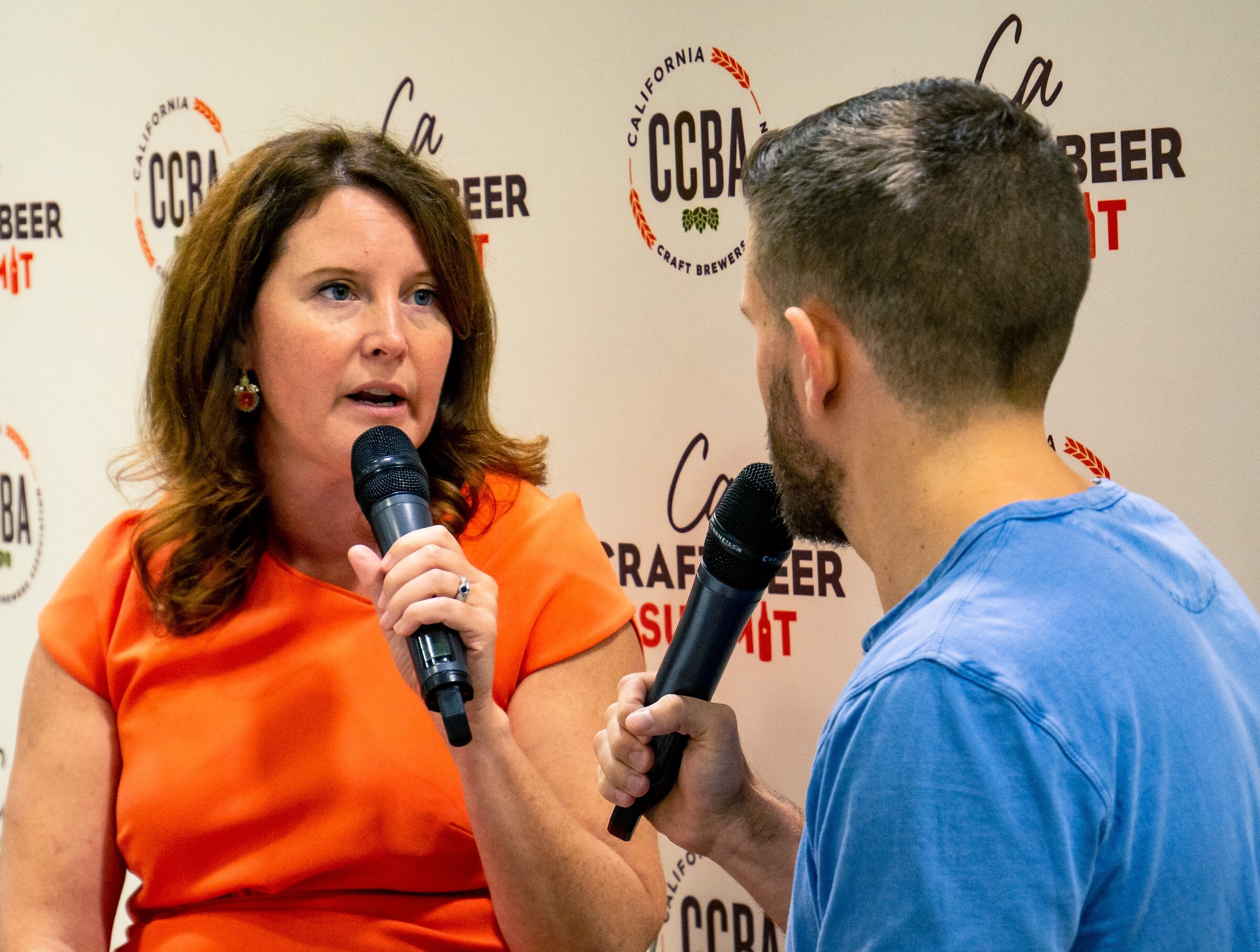 A woman with shoulder-length brown hair in an orange dress holds a microphone in front of the CCBA backdrop, facing an interviewer in a blue T-shirt.