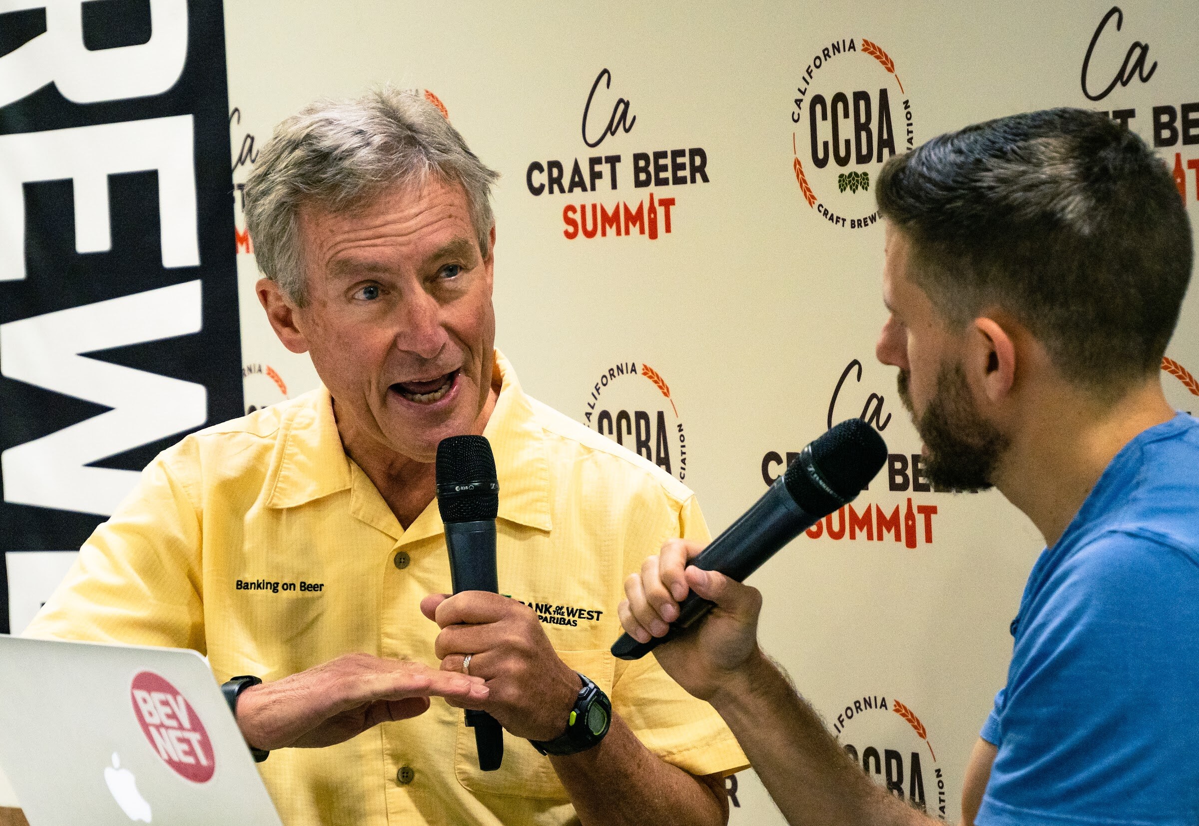 A gray-haired man in a yellow shirt reading Banking on Beer and Bank of the West gestures with a microphone in front of a stickered laptop.