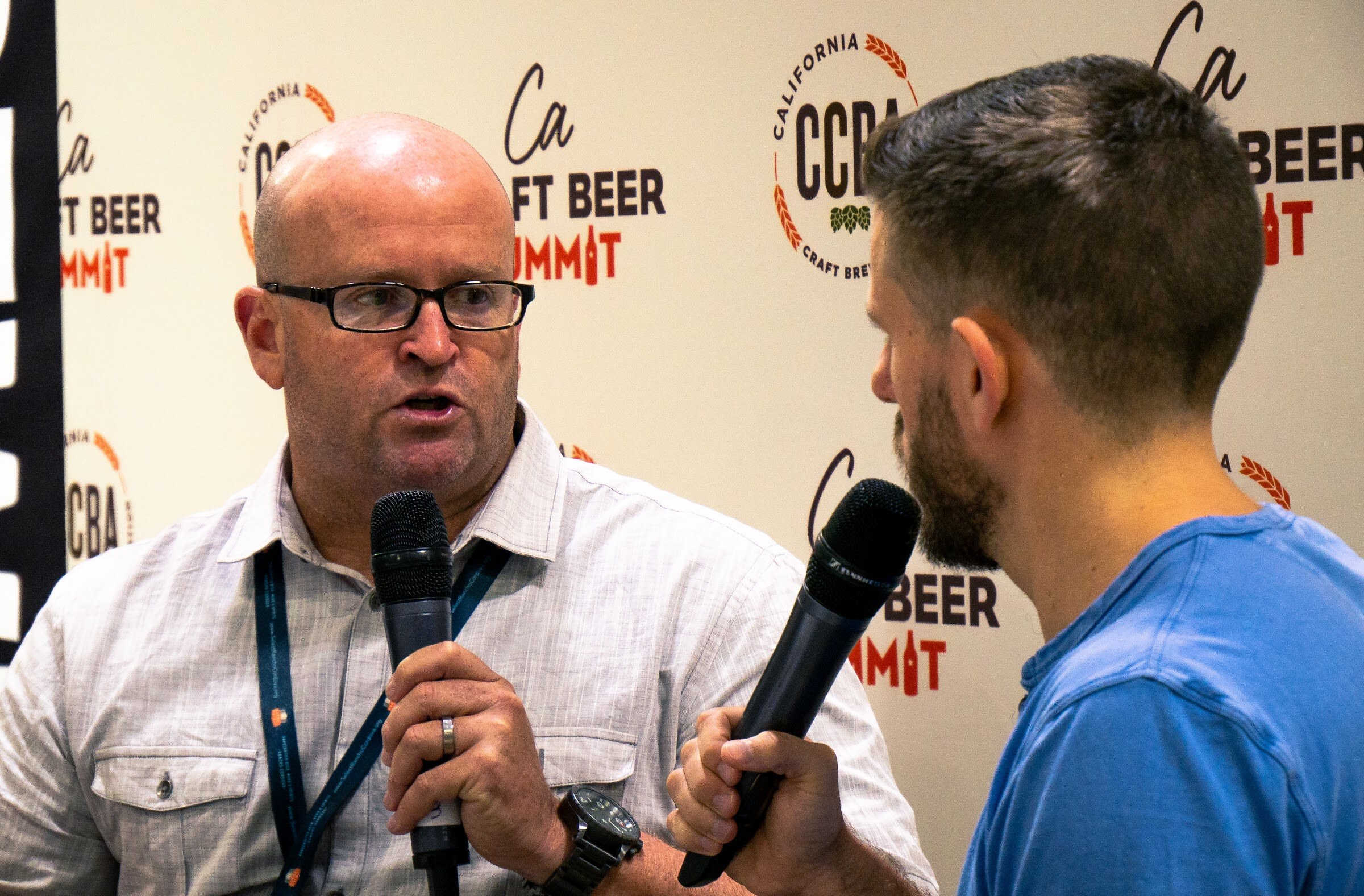 A bald man in glasses and a gray button-down shirt speaks into a microphone in front of the CCBA backdrop, facing an interviewer in a blue T-shirt.