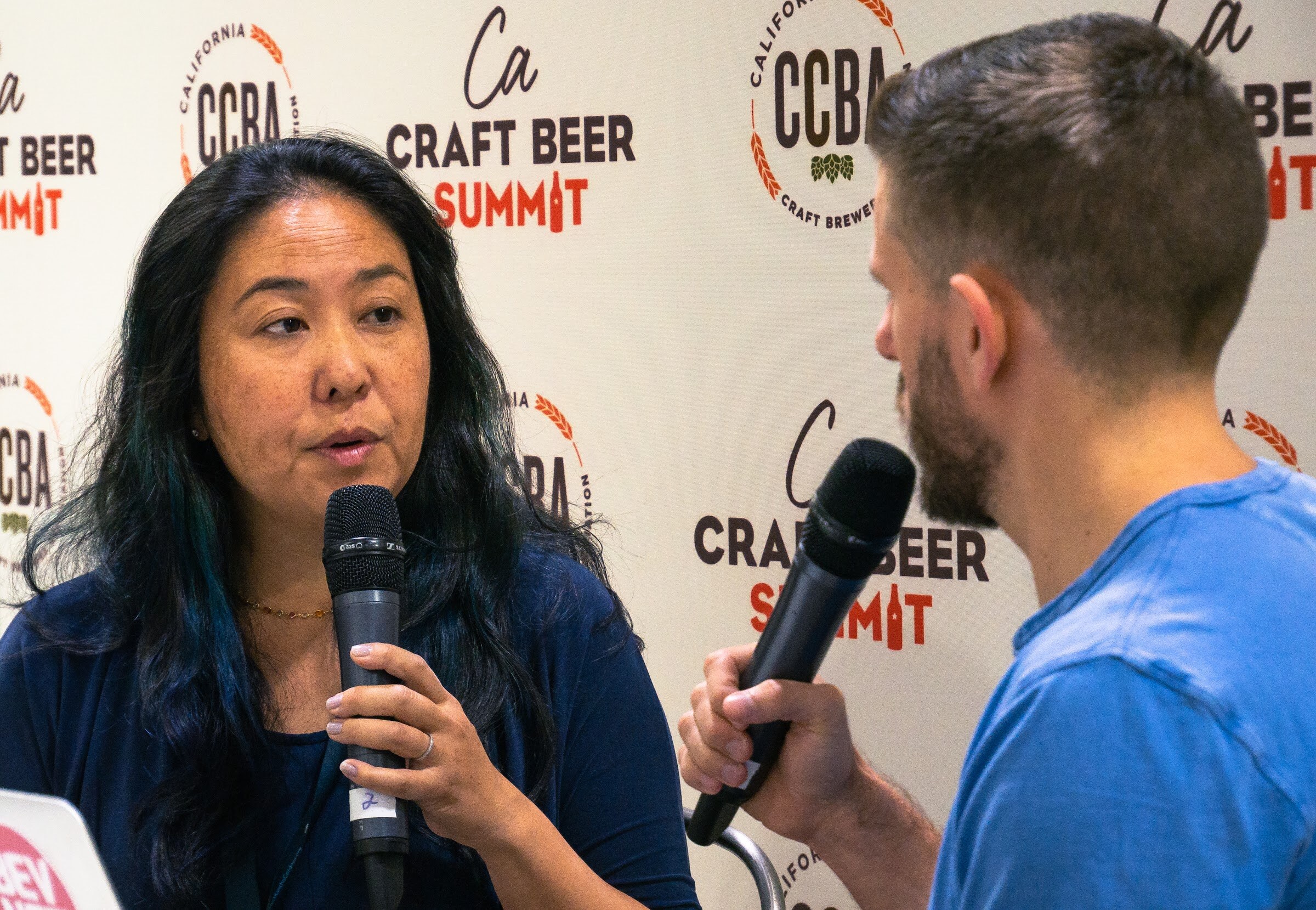 A woman with long dark hair and a navy top holds a microphone, with part of a BevNET laptop sticker visible at lower left.