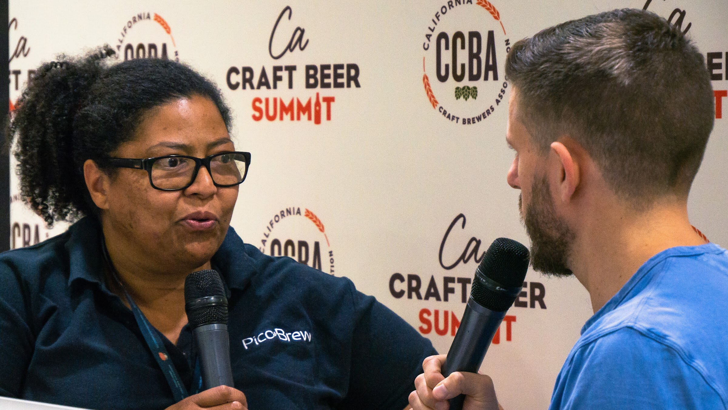 A woman in glasses and a navy PicoBrew polo holds a microphone in front of the CCBA backdrop, with the interviewer in a blue T-shirt at right.