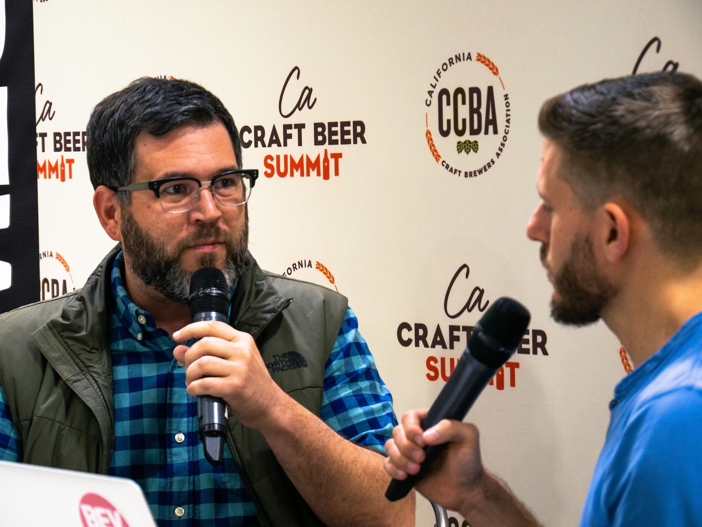 A bearded man in glasses, a teal plaid shirt and an olive vest holds a microphone in front of a CCBA backdrop, with the edge of a BevNET laptop at lower left.