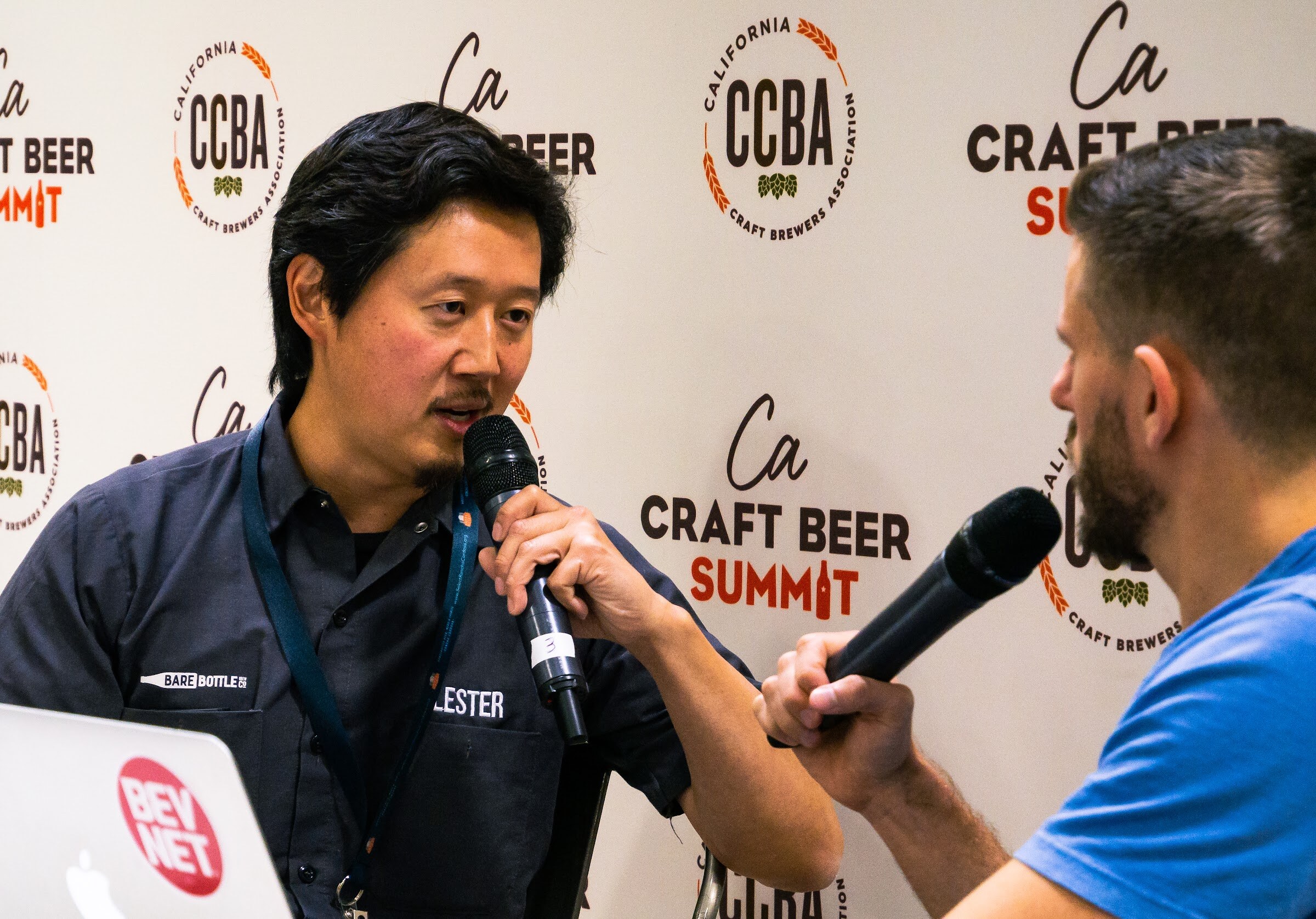 A man in a dark work shirt with a Bare Bottle logo and a lanyard holds a microphone beside a laptop with a BevNET sticker.