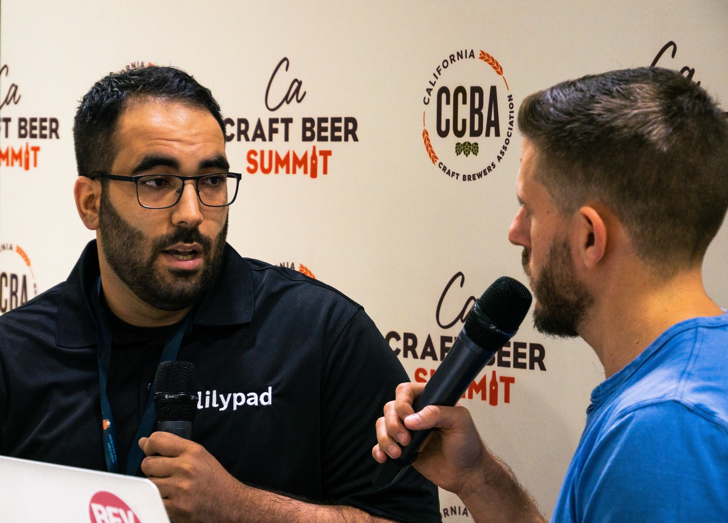 A bearded man in glasses and a black polo with a Lilypad logo holds a microphone in front of the CCBA backdrop, facing the interviewer.