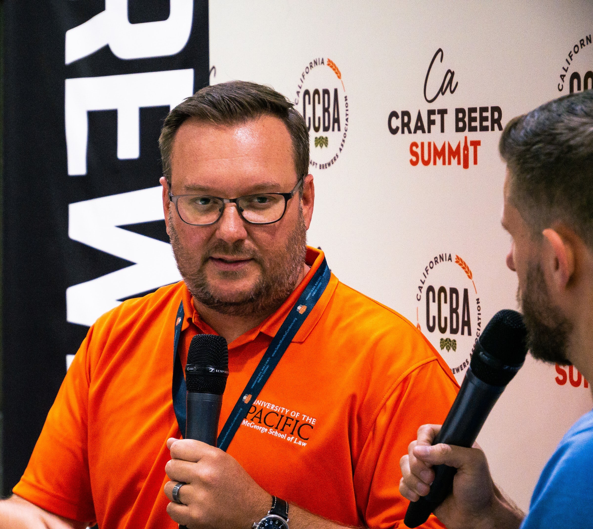 A man in glasses and an orange polo with a School of Law logo and a lanyard holds a microphone in front of a Brewbound banner and the CCBA backdrop.