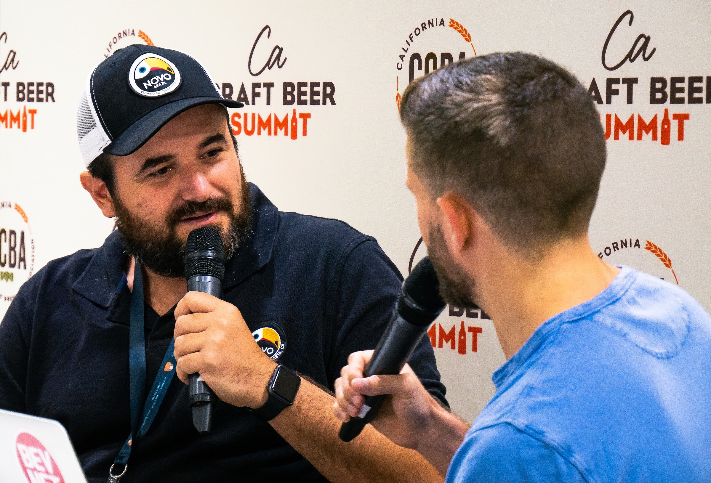 A bearded man in a black Novo Brazil trucker cap and polo with a lanyard speaks into a microphone, with a BevNET laptop at lower left.