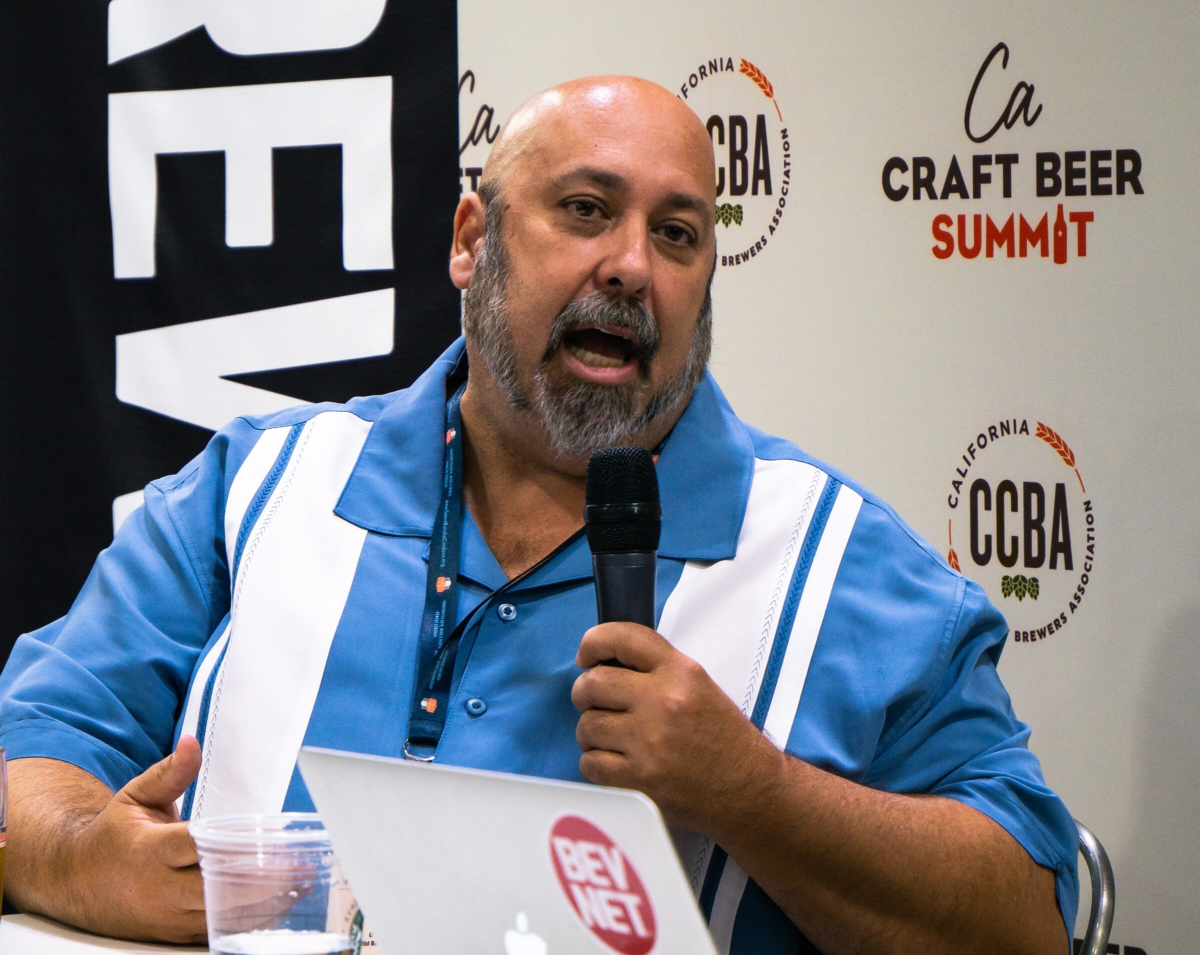 A bald, gray-bearded man in a blue and white shirt with a lanyard speaks into a microphone, with a BevNET laptop and a plastic cup on the table.