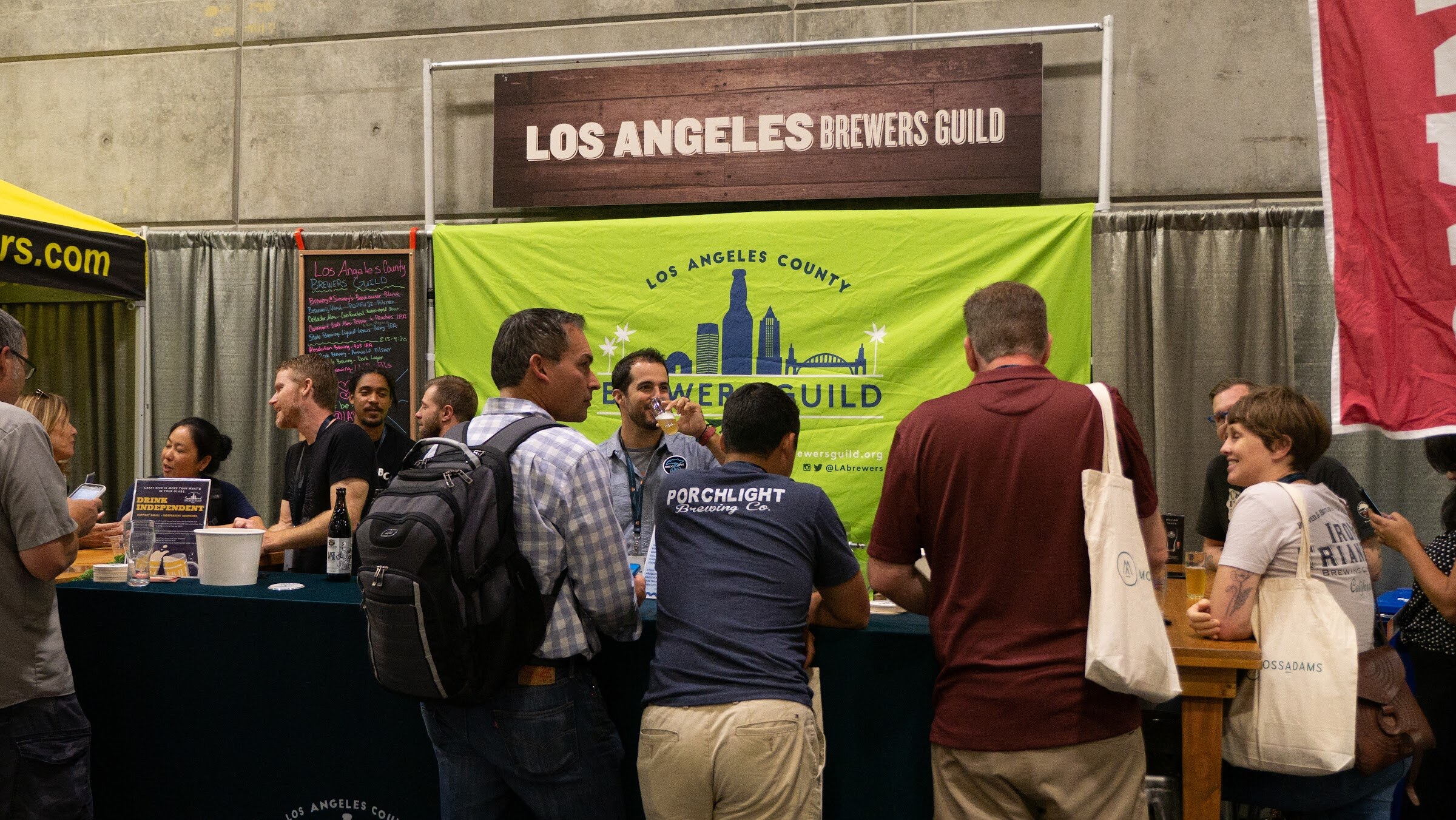 Several attendees, one in a Porchlight Brewing shirt, stand at a counter in front of a green banner and a wooden sign reading Los Angeles Brewers Guild.