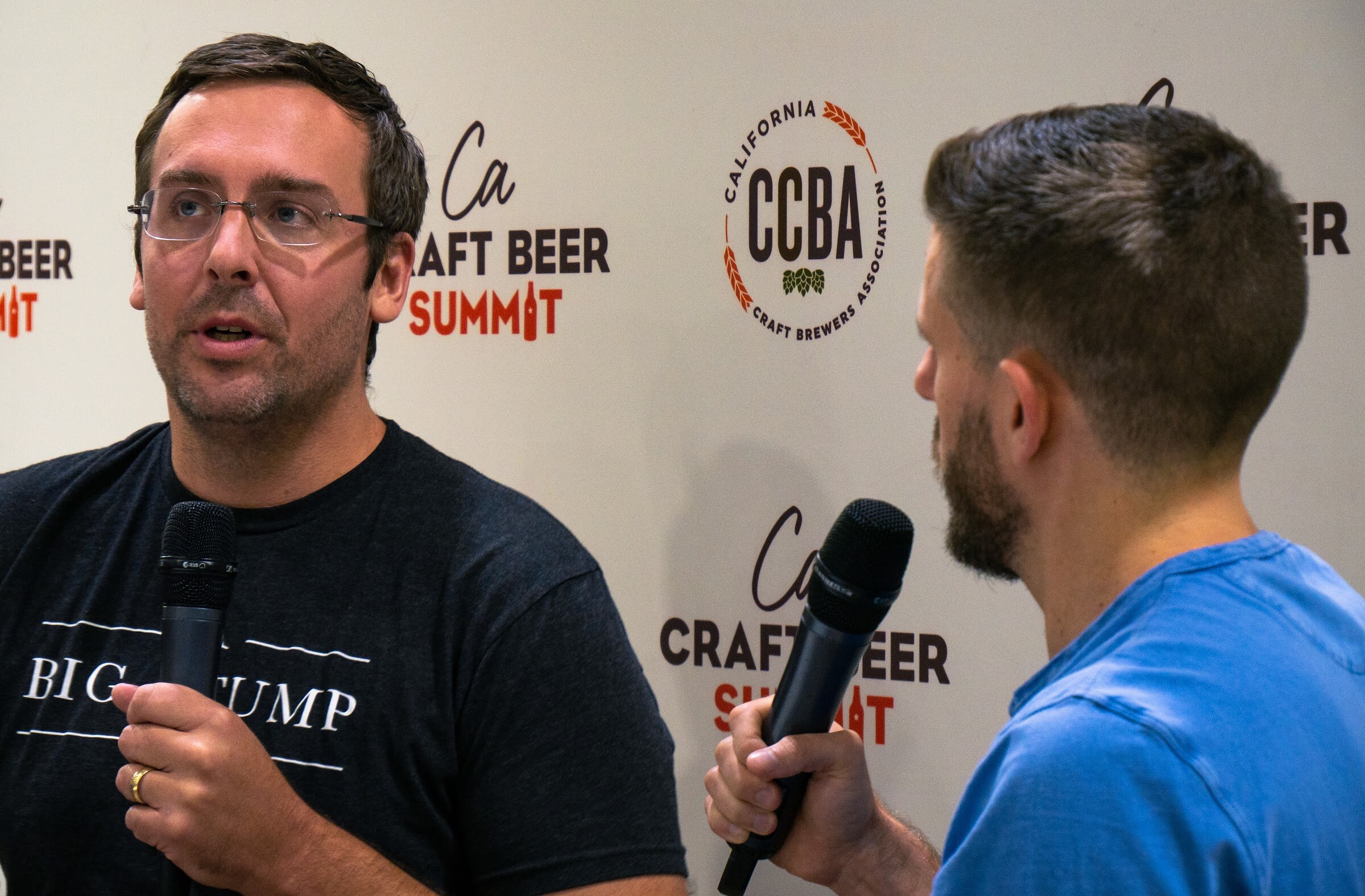 A man in glasses and a dark Big Stump T-shirt holds a microphone in front of the CCBA backdrop, with the interviewer seen from behind.