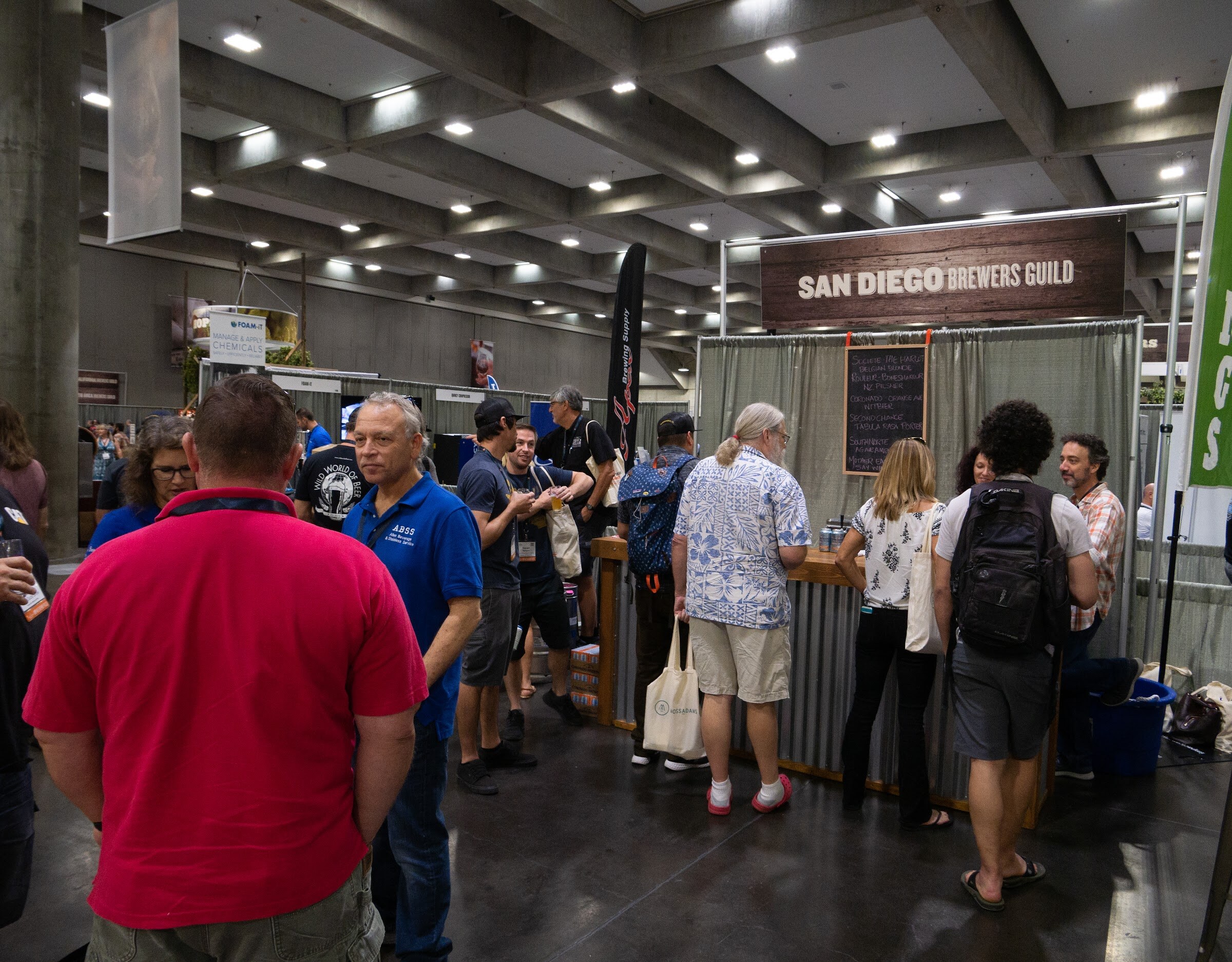 A busy aisle in a concrete convention hall with attendees at a corrugated bar beneath a San Diego Brewers Guild sign and a chalkboard menu.