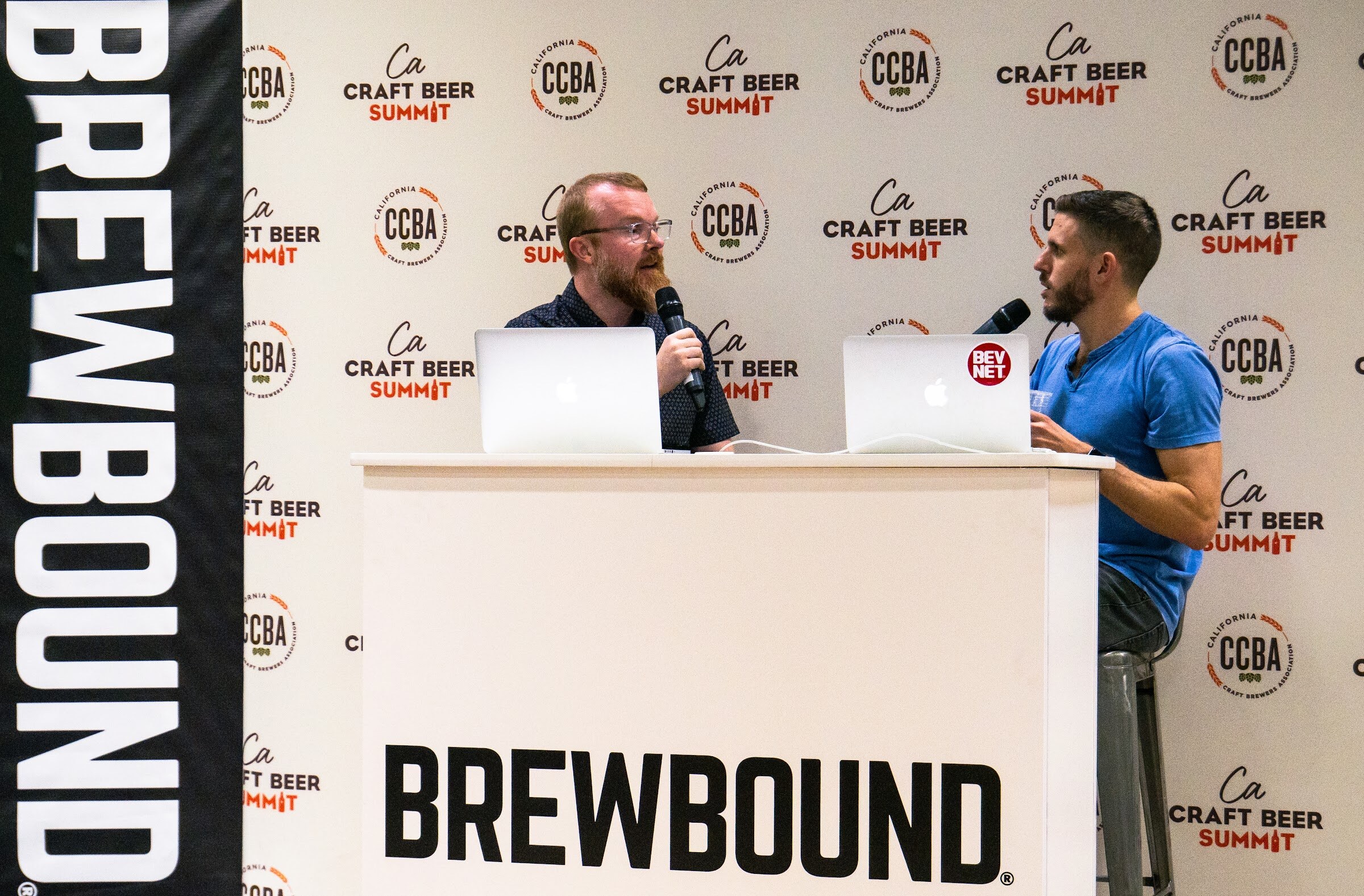 Two men at a white Brewbound podium, one in glasses and a dark shirt and one in a blue T-shirt, speaking into microphones in front of the California Craft Beer Summit backdrop and a Brewbound banner.