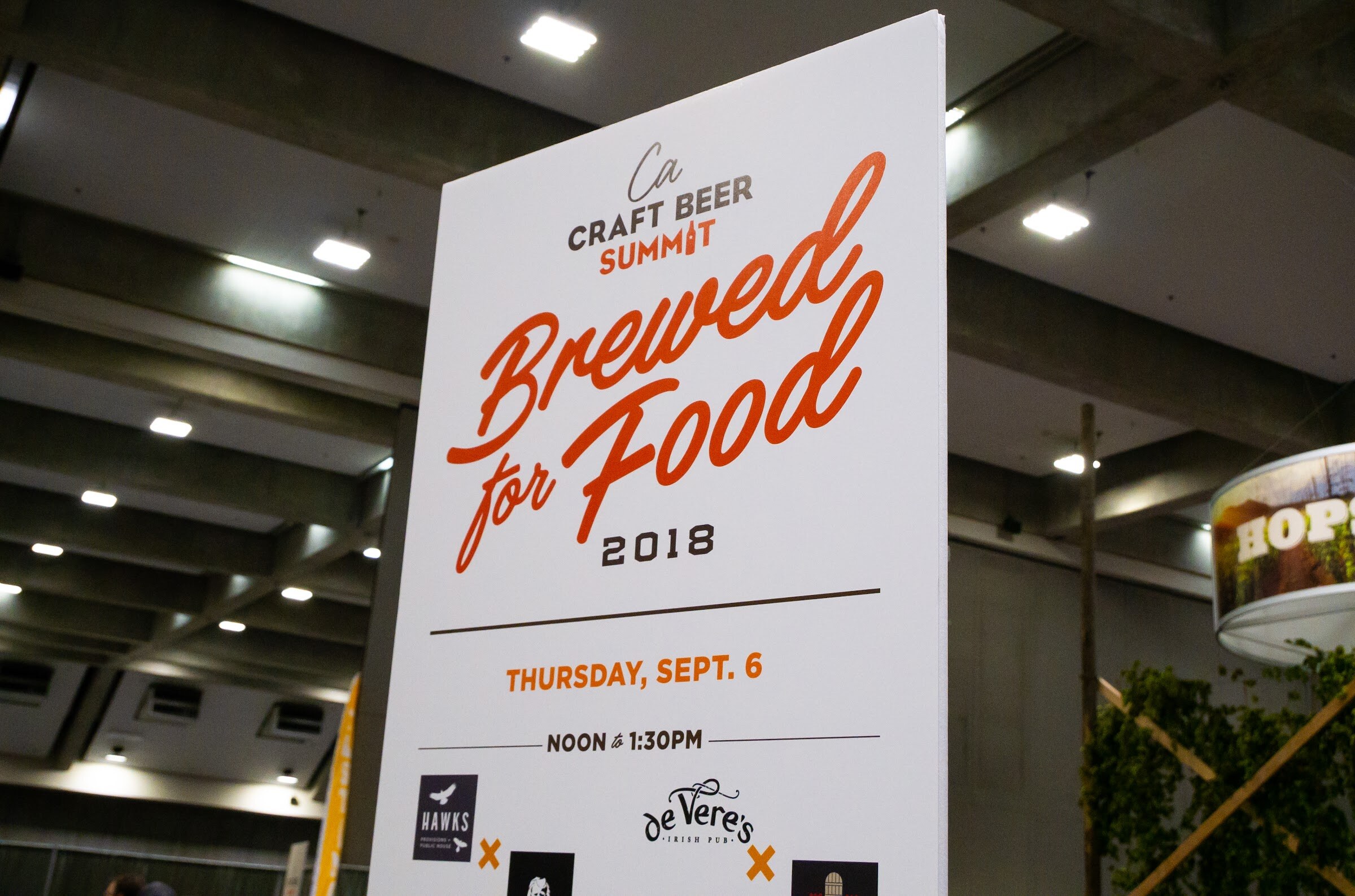 A tall white sign reading Ca Craft Beer Summit Brewed for Food 2018, Thursday Sept. 6, noon to 1:30 PM, with restaurant logos below, in a concrete exhibit hall.