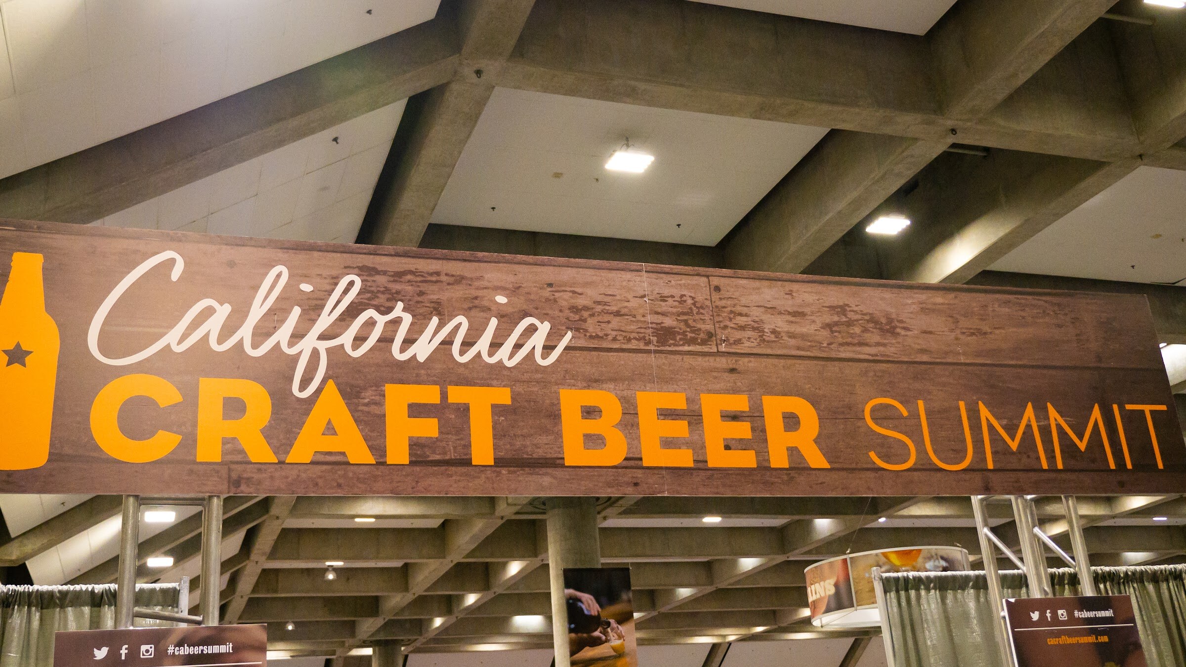 A wood-grain banner reading California Craft Beer Summit in white and orange letters, hanging from the concrete ceiling grid of the exhibit hall.
