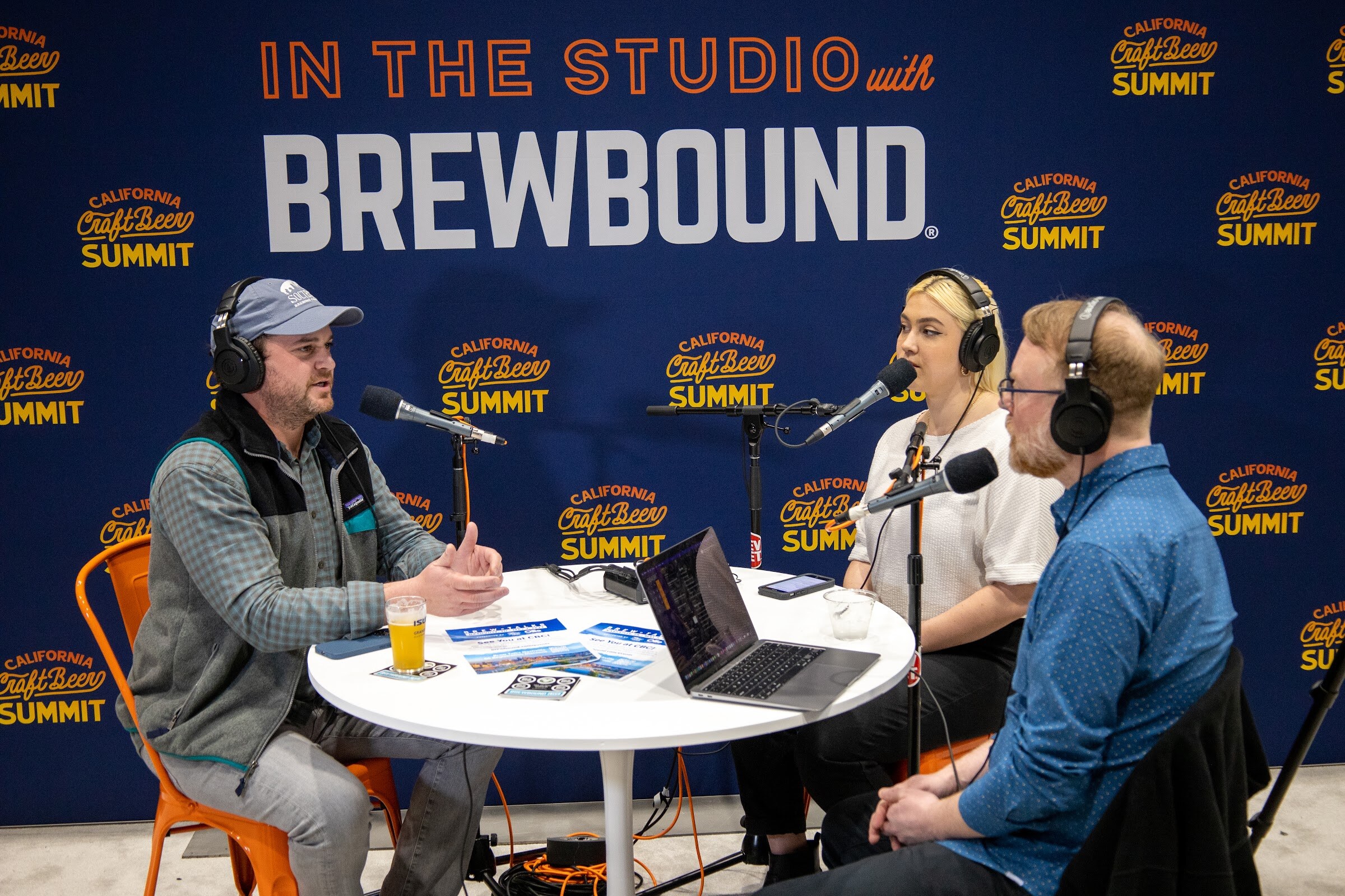 Three people in headphones talk at a round table with a beer glass and a laptop in front of an In the Studio with Brewbound and California Craft Beer Summit backdrop