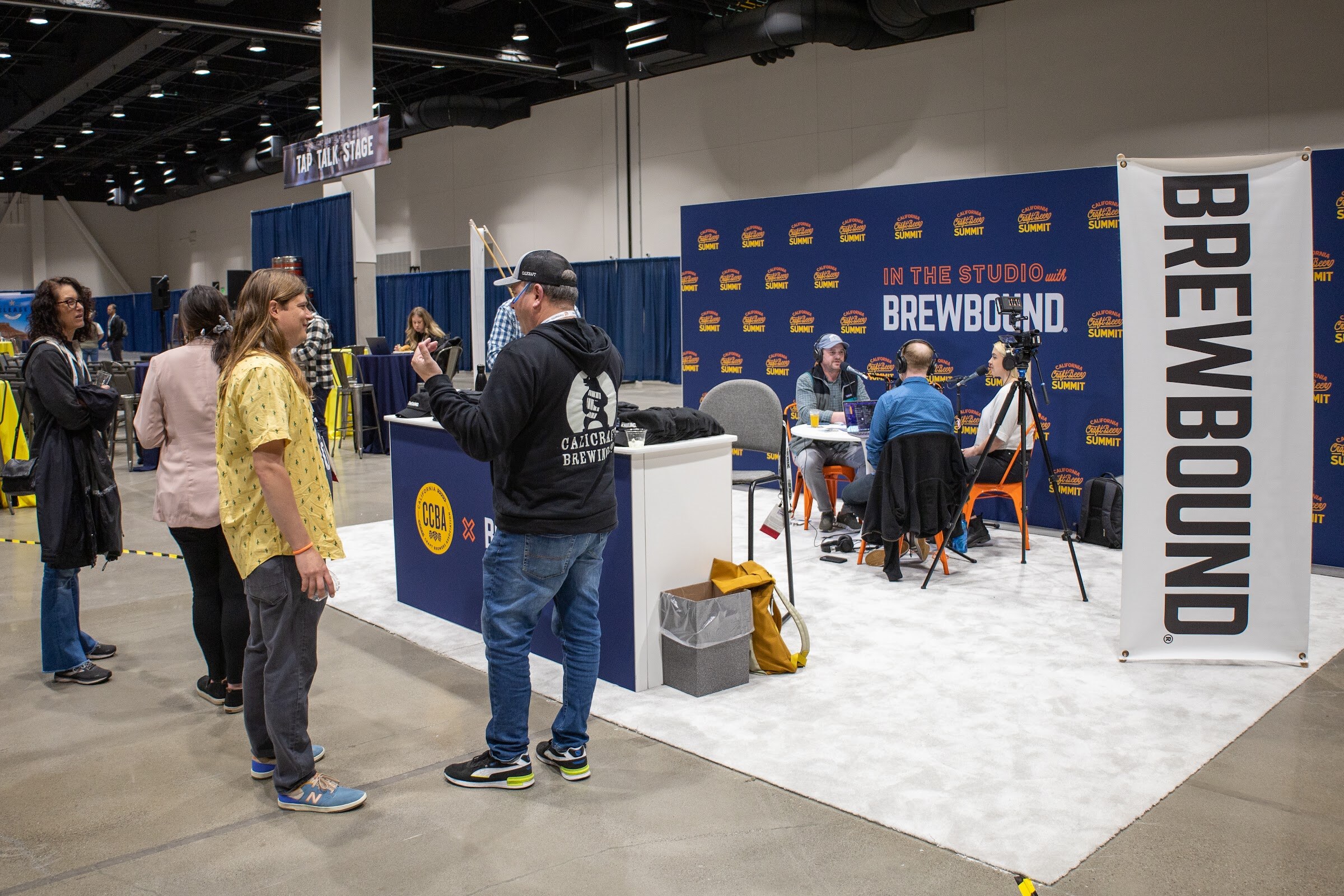A wide view of the Brewbound podcast booth with a navy backdrop and a tall Brewbound banner while attendees stand and talk nearby on the expo floor