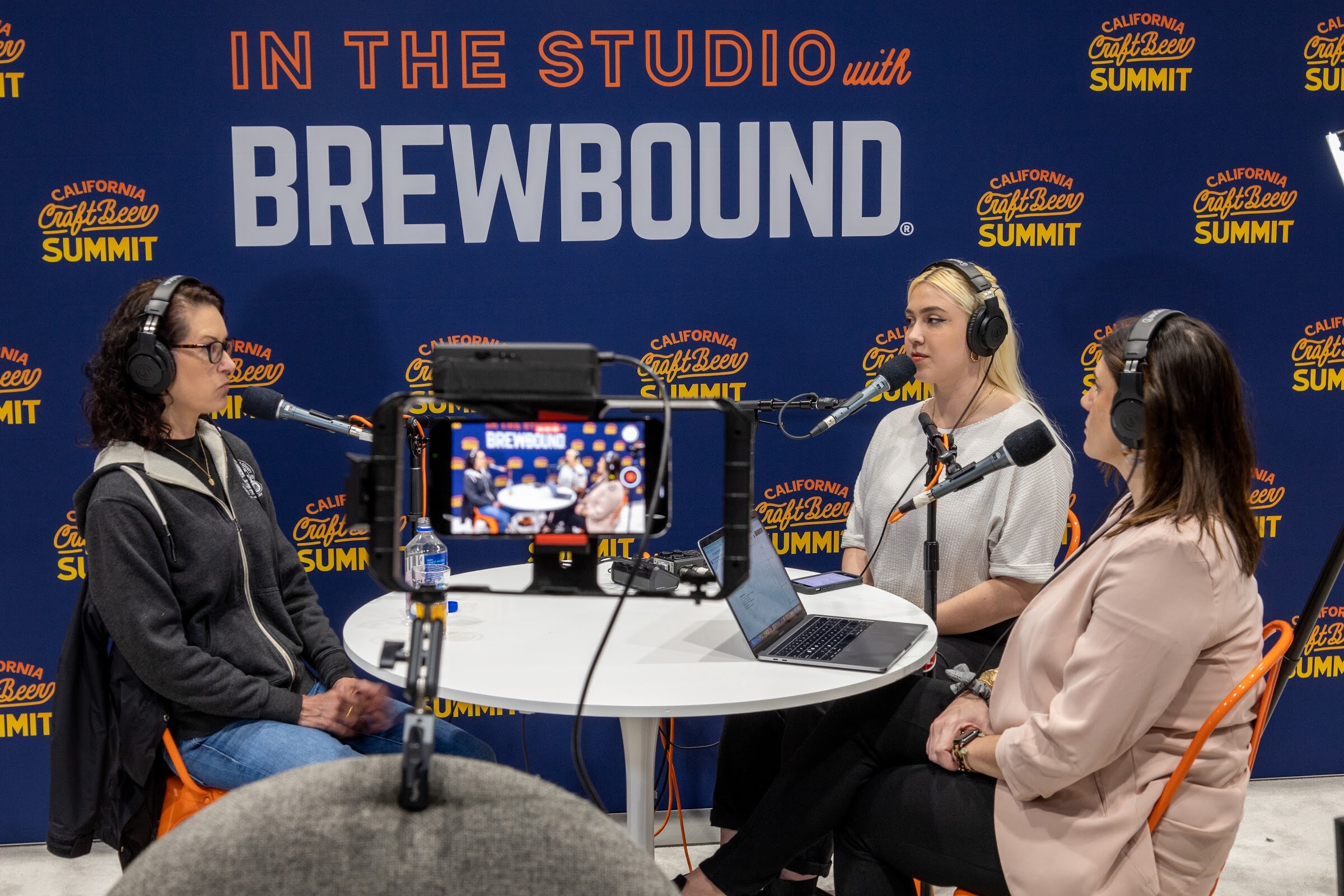 Three people in headphones sit around a white table with a laptop and a phone on a camera rig in front of the In the Studio with Brewbound backdrop