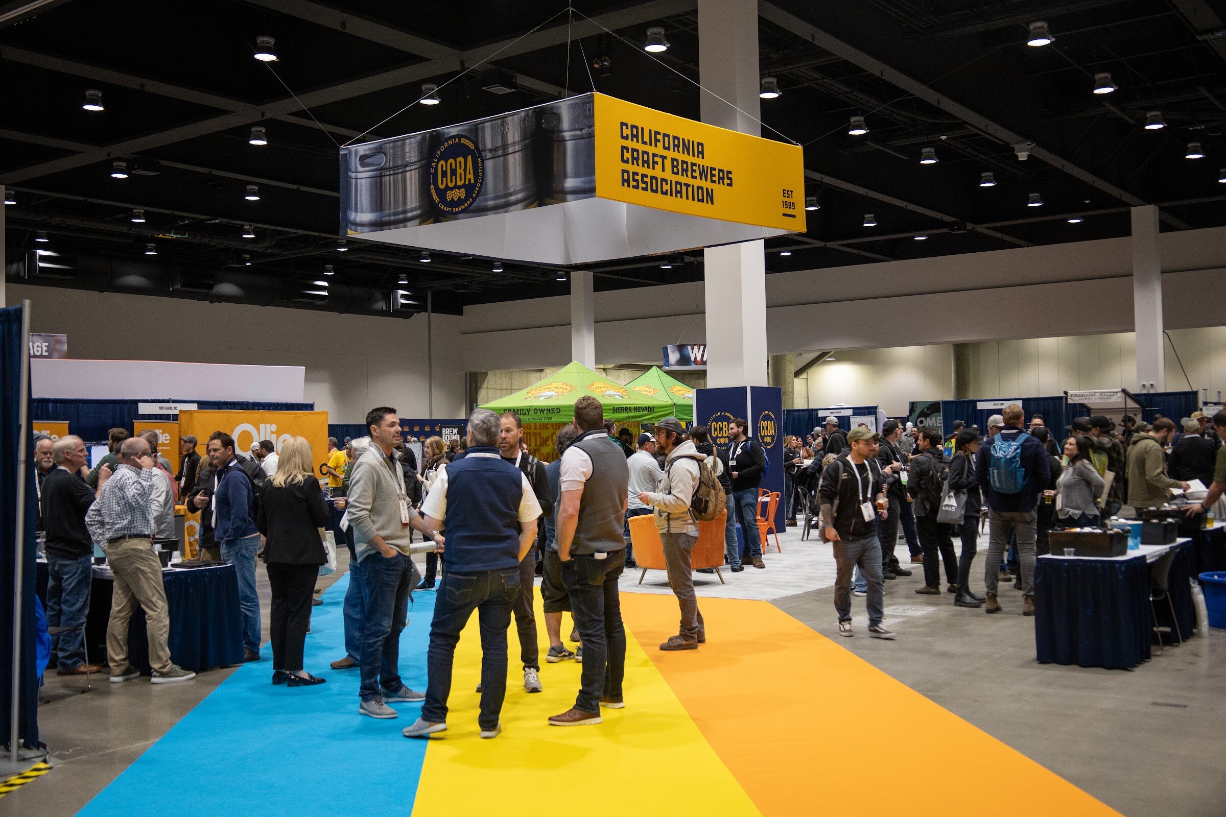 A hanging yellow California Craft Brewers Association sign above crowds of attendees walking along a colorful striped aisle on the expo floor